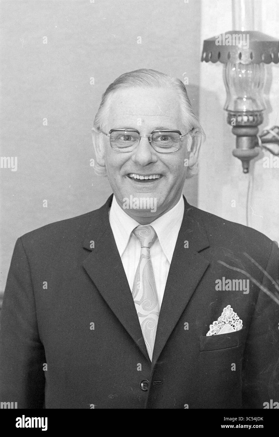 Porträt von Mr. More, Portraits, 25-04-1974 Whizgle News, Dutch Desk, Niederlande, 1950-2000 Ein lächelnder Mann in einem Anzug mit gemusterter Krawatte und einem Taschenfeld steht neben einer kunstvollen Lampe und strahlt ein freundliches und nahes Auftreten aus. Stockfoto