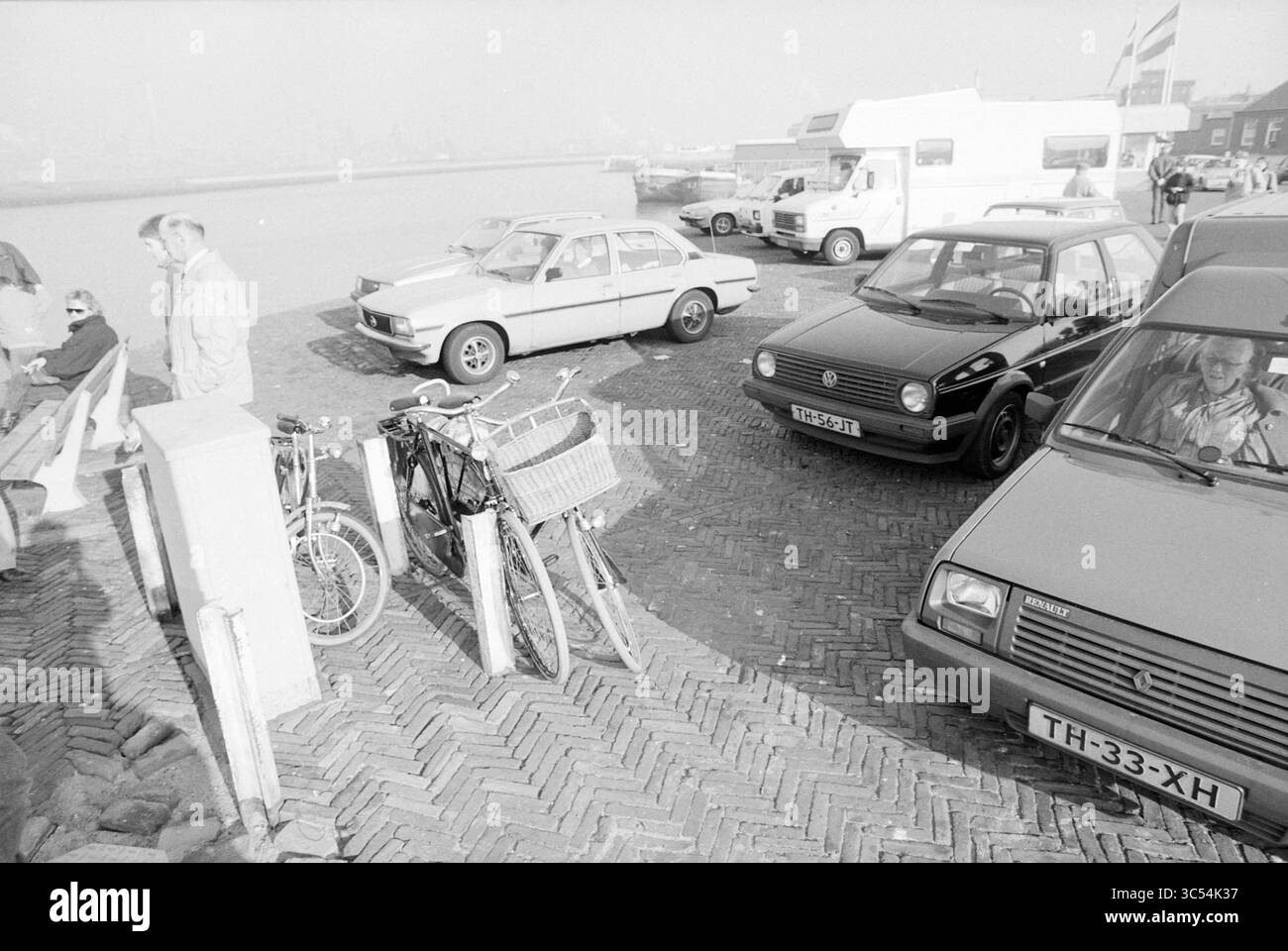 Geschäftiger Leiter des Hafens, Ports, 08-02-1989 Whizgle News, Dutch Desk, Niederlande, 1950-2000 Eine geschäftige Szene in einem Hafen, in dem mehrere Autos neben Fahrrädern parken und eine Mischung aus Fahrzeugen zeigen, die typisch für eine frühe Zeit sind. Die Menschen genießen die Atmosphäre gemütlich, wobei eine Person auf einer niedrigen Mauer sitzt, während andere in der Nähe arbeiten. Die Umgebung ist lebhaft und deutet auf einen anstrengenden Tag am Wasser hin. Stockfoto