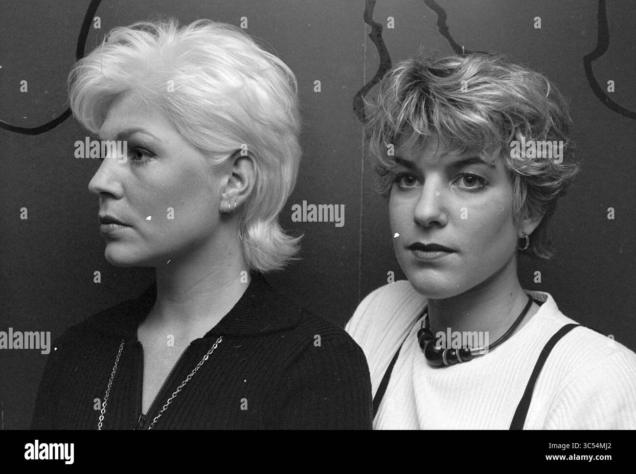 Two Haircuts, Loni Korver, 02-03-1995 Whizgle News, Dutch Desk, Niederlande, 1950-2000 zwei Frauen stehen nebeneinander und zeigen jeweils unterschiedliche Frisuren und Ausdrucksformen. Eine hat kurzes, schlankes platinblondes Haar, während die andere mit zerzausten, welligen Haaren ausgestattet ist. Ihr kontrastierender Look erinnert an verschiedene Stile und Persönlichkeiten, eingerahmt vor einem schlichten Hintergrund. Stockfoto