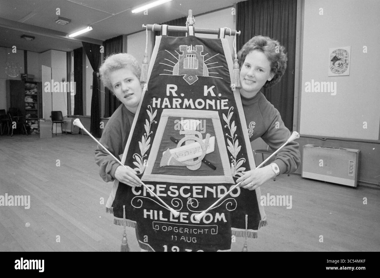 2 Majorette-Musikband Crescendo, Music, People, 19-04-1988 Whizgle News, Dutch Desk, Niederlande, 1950-2000 zwei Personen halten ein dekoratives Banner mit dem Emblem von „Crescendo“ und dem Jahr 1973, das ihre Teilnahme an einer Musikgruppe oder Veranstaltung zeigt. Sie posieren spielerisch mit dem Banner, das mit verschiedenen künstlerischen Elementen verziert ist, in einem Raum mit Vorhängen und Holzboden. Stockfoto
