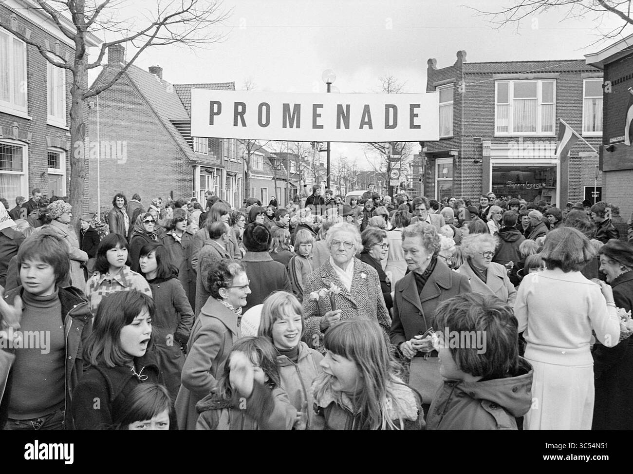Promenade in Lisse, 07-04-1977 Whizgle News, Dutch Desk, Niederlande, 1950-2000 Eine geschäftige Menschenmenge versammelt sich in einer lebendigen Straßenszene, wo ein großes Schild mit der Aufschrift „PROMENADE“ darüber hängt. Menschen jeden Alters führen Gespräche, einige lächeln, während andere tief in Gedanken erscheinen, wodurch eine Atmosphäre der Gemeinschaft und der Vorfreude entsteht. Von Bäumen gesäumte Gebäude säumen die Gegend und tragen zur lebhaften Energie der Versammlung bei. Stockfoto