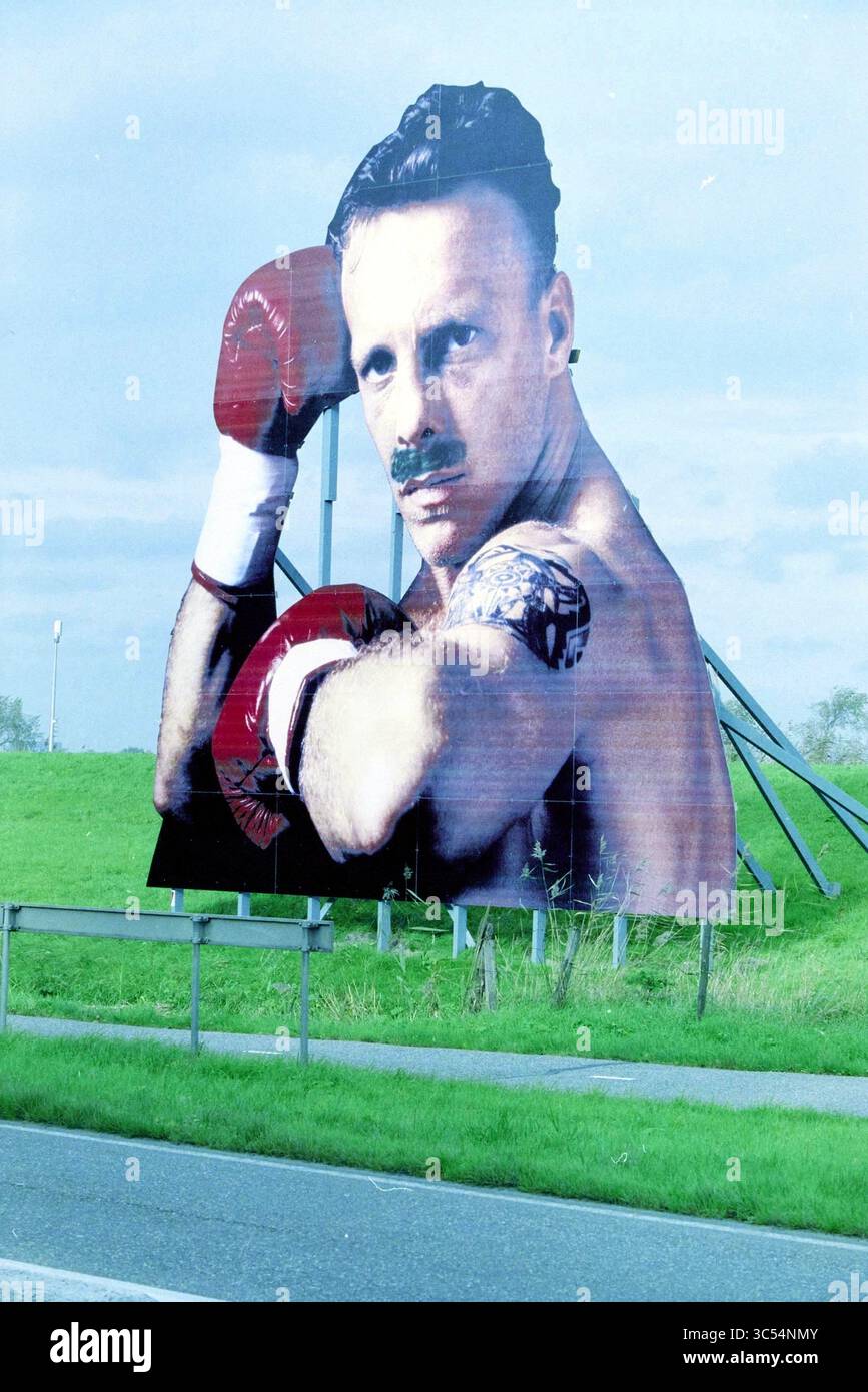 Billboard Amsterdamseweg, IJmuiden, IJmuiden, Niederlande, 28-10-2001 Whizgle News, Dutch Desk, Niederlande, 1950-2000 Eine große Plakatwand am Straßenrand zeigt einen muskulösen Boxer mit einem markanten Schnurrbart, der rote Boxhandschuhe trägt und eine heftige Pose zeigt. Der Hintergrund von üppigem grünem Gras verstärkt die beeindruckende visuelle Wirkung der Werbung. Stockfoto