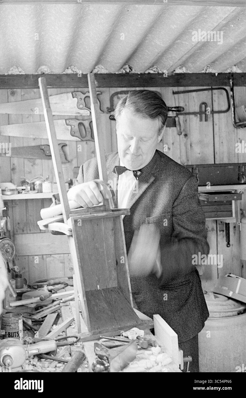 Prof. v.d. Mountain, Persons, 02-07-1965 Whizgle News, Dutch Desk, Niederlande, 1950-2000 Ein fokussierter Handwerker arbeitet intensiv an einem Holzprojekt, umgeben von Werkzeugen und Holzbearbeitungsmaterialien in einer Werkstatt. Er passt ein Möbelstück an und zeigt sein Können und seine Hingabe an sein Handwerk. Stockfoto