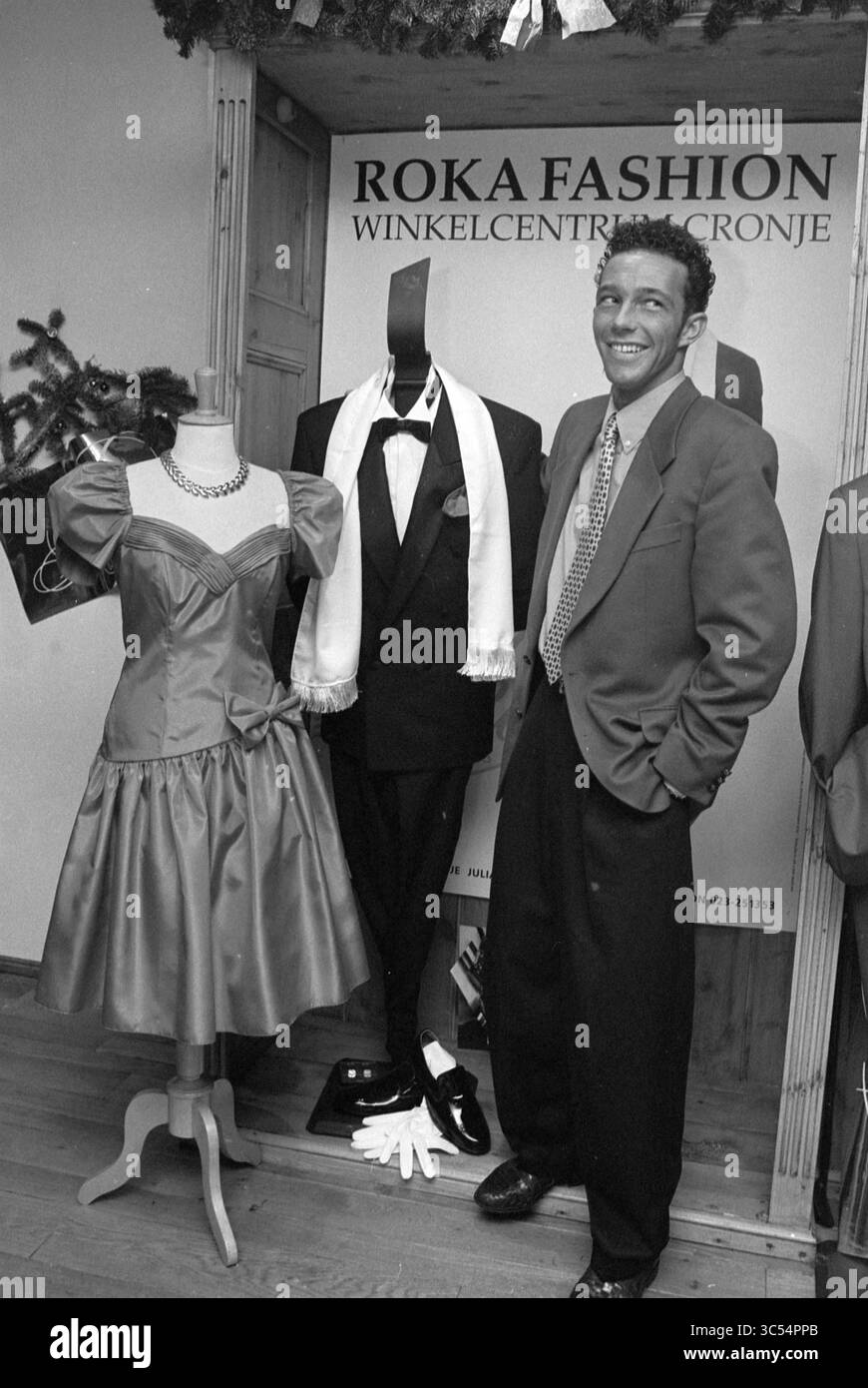 Partykleidung, Roka Fashion, Haarlem, Niederlande, 19-12-1993 Whizgle News, Dutch Desk, Niederlande, 1950-2000 Ein stilvolles Boutique-Display zeigt ein elegantes Ballkleid neben einem klassischen Smoking, während ein lächelndes Individuum selbstbewusst posiert und einen modischen Look zeigt. Die Atmosphäre ist fröhlich und lebendig, spiegelt ein Gefühl von Feier und Stil wider. Stockfoto