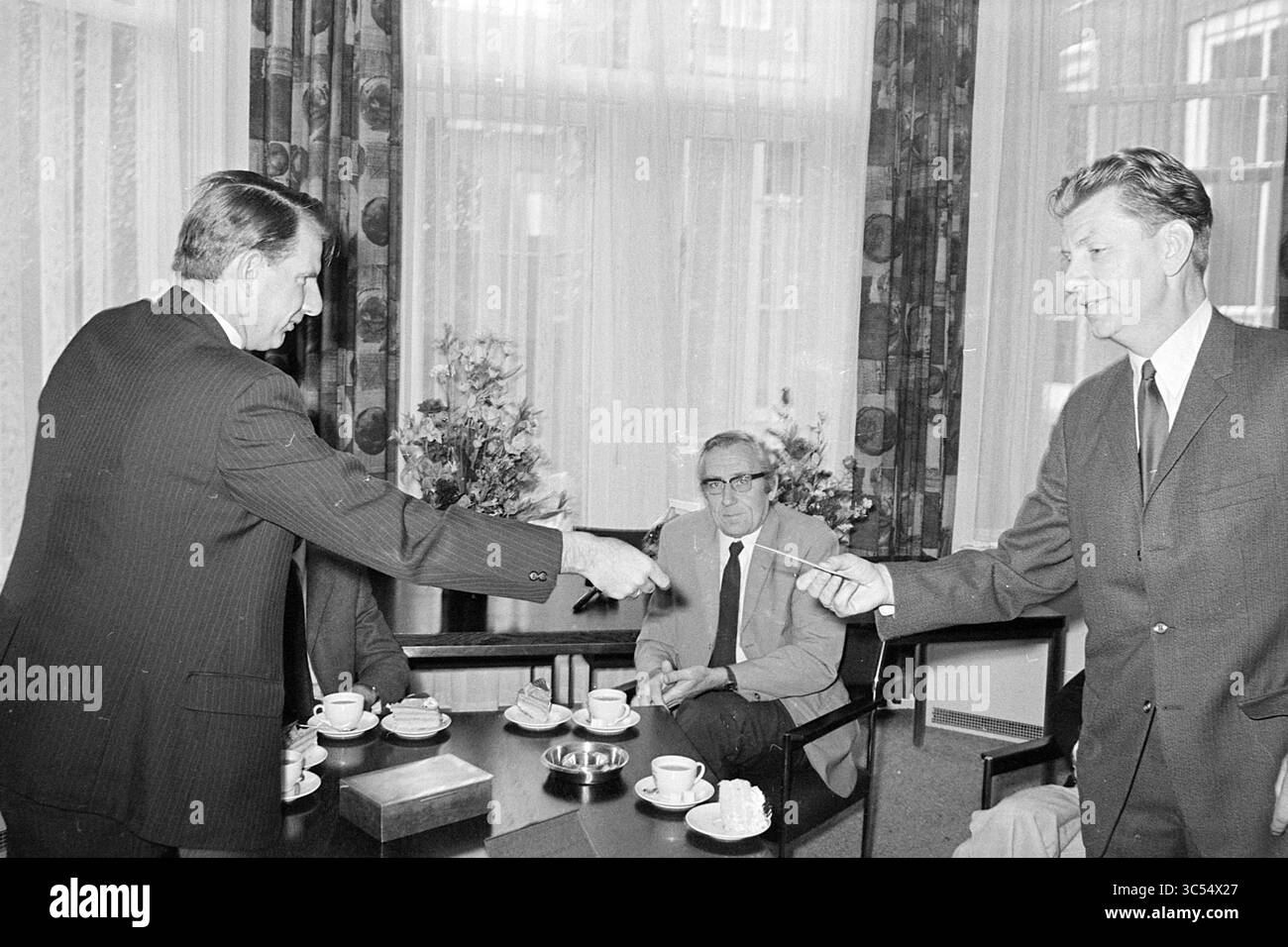 Ehren von 3 Schullehrern., Ehren, 23-06-1972 Whizgle News, Dutch Desk, Niederlande, 1950-2000 zwei Männer in Anzügen haben einen Handschlag, der ein bedeutendes Abkommen oder eine Partnerschaft symbolisiert. Ein dritter Mann sitzt in der Nähe und beobachtet die Interaktion, während ein Tisch mit Kaffeetassen und einem kleinen Dessert gedeckt ist. Die Einrichtung verfügt über große Fenster mit Vorhängen und Blumenarrangements, die eine formelle Atmosphäre vermitteln. Stockfoto