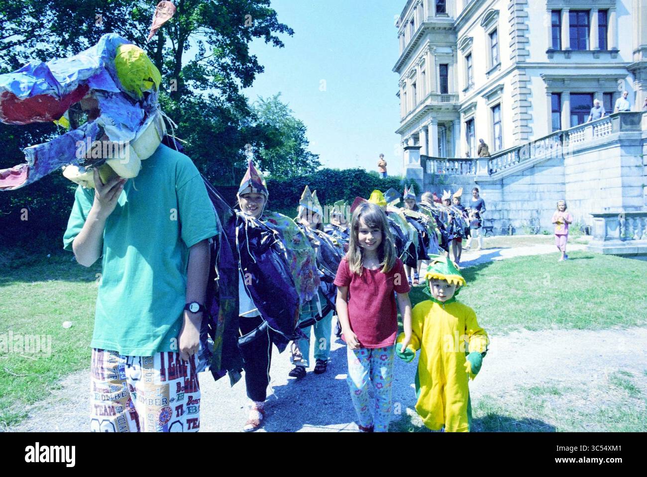 Kinderschach, Elswout, 13-07-1995 Whizgle News, Dutch Desk, Niederlande, 1950-2000 Eine Gruppe von Kindern in bunten Kostümen zieht fröhlich in einer Schlange, wobei ein Kind eine Drachenmaske trägt. Sie befinden sich in einer üppigen Umgebung im Freien, in der Nähe eines beeindruckenden Gebäudes, was eine festliche Atmosphäre schafft. Stockfoto