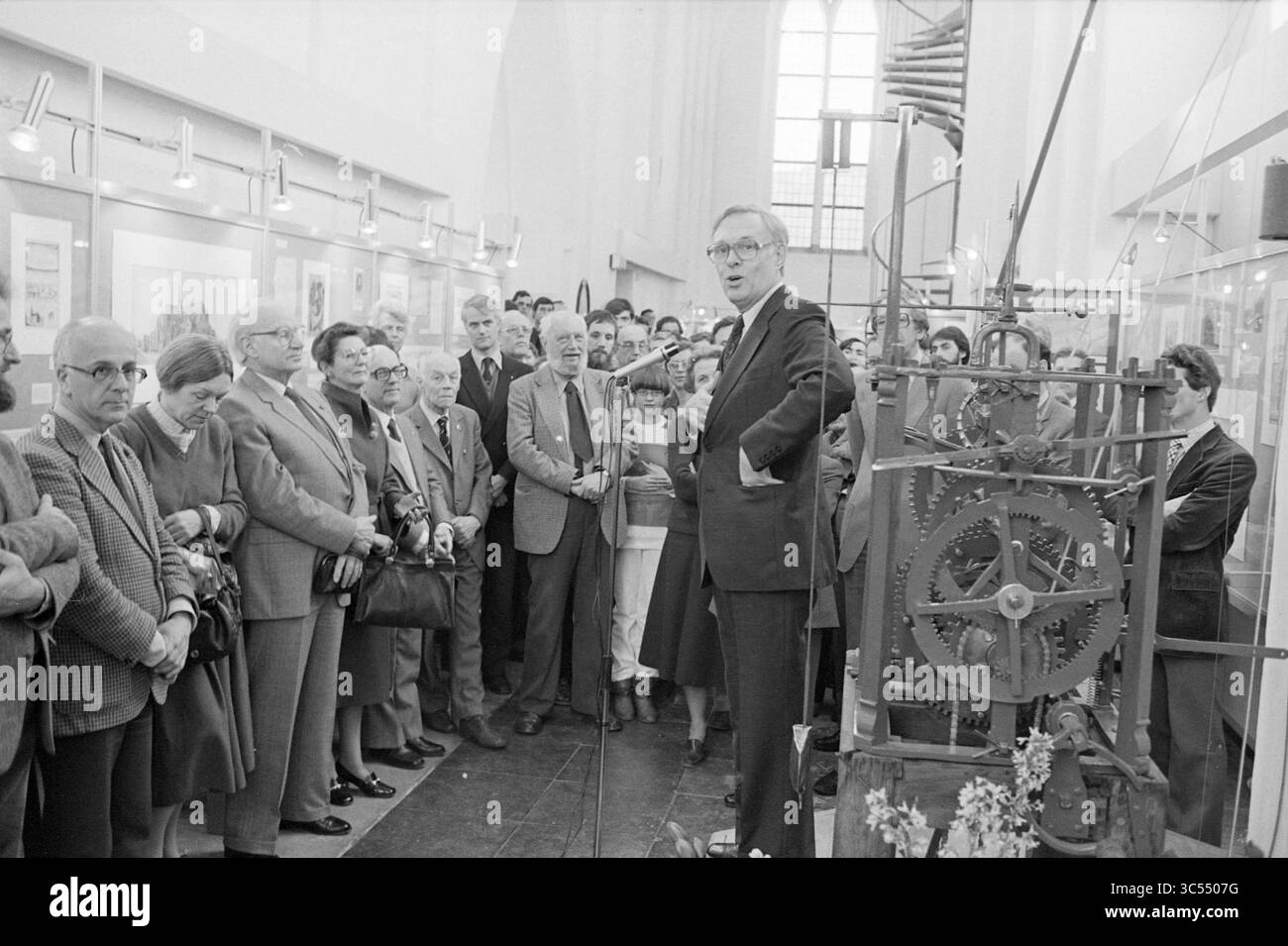 Eröffnung der Ausstellung "das Archiv: Haarlem Gedächtnis", anlässlich 125 Jahre Stadtarchiv, durch Bürgermeister Reehorst, in der Janskerk., Jahrestag, Jahrestag, Haarlem, Jansstraat 40, Niederlande, 25-03-1982 Whizgle News, Dutch Desk, Niederlande, 1950-2000 Eine Gruppe von Menschen hört während einer Veranstaltung aufmerksam einem Sprecher zu, der in der Nähe einer großen, aufwändig gestalteten mechanischen Uhr steht. Das Publikum ist vielfältig, darunter Männer und Frauen, und sie scheinen engagiert und neugierig auf die Präsentation und das historische Artefakt zu sein. Stockfoto