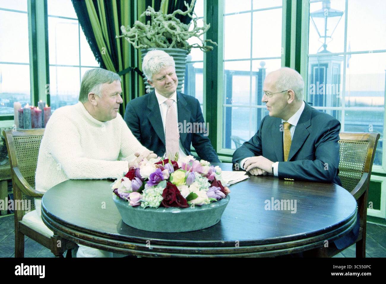 H. Mens, H. Oljans, Zalm Noordwijk, Noordwijk, 10-01-2003 Whizgle News, Dutch Desk, Niederlande, 1950-2000 drei Männer unterhalten sich an einem runden Tisch, der mit einem floralen Mittelstück geschmückt ist. Die Umgebung verfügt über große Fenster, die einen Blick nach außen bieten, und die allgemeine Atmosphäre lässt ein Treffen von Kollegen in einem beruflichen oder sozialen Kontext vermuten. Stockfoto