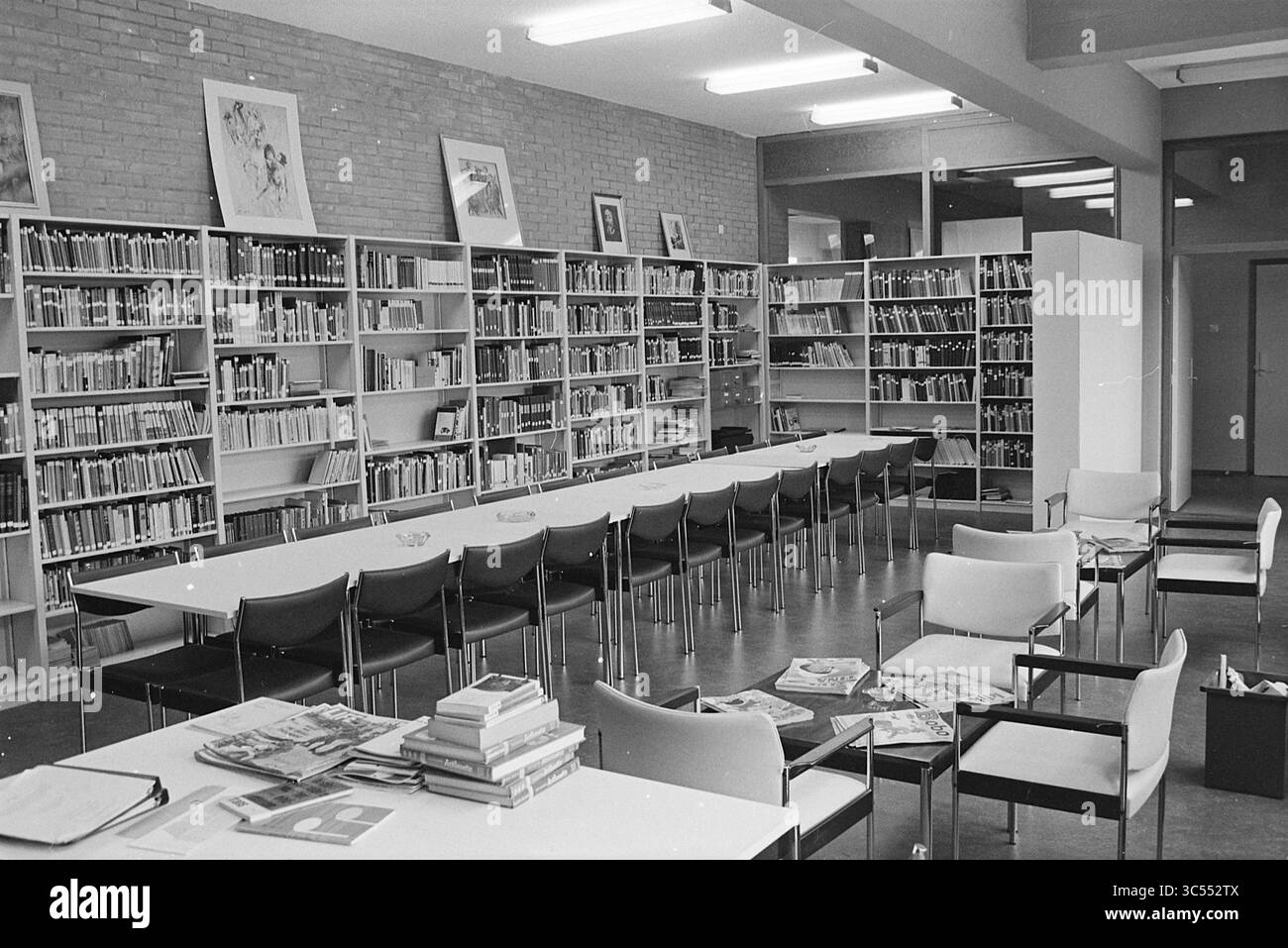B.C.-Kindergarten Santpoort, Schulen, Santpoort-Zuid, Bloemendaalsestraatweg, 14-08-1969 Whizgle News, Dutch Desk, Niederlande, 1950-2000 Eine geräumige Bibliothek mit hohen Regalen mit Büchern, ordentlich organisiert, und einem langen Tisch, umgeben von Stühlen in der Mitte. Über einen anderen Tisch sind Magazinstapel verteilt, und große Fenster lassen den Untersuchungsbereich durch natürliches Licht ausleuchten. Wandbilder verleihen der Umgebung einen Hauch von Kreativität. Stockfoto