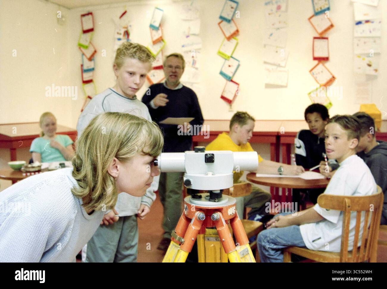Survey, Hoofddorp, Niederlande, 10-06-2002 Whizgle News, Dutch Desk, Niederlande, 1950-2000 Eine Gruppe von Kindern versammelt sich in einem Klassenzimmer, von denen einige an Schreibtischen sitzen, während andere sich auf ein Vermessungsinstrument konzentrieren. Ein Mädchen blickt in das Gerät und zeigt Neugier und Engagement, während ein erwachsener Lehrer Anleitung gibt. Die Wände sind mit farbenfrohen Kunstwerken geschmückt, die von den Schülern gefertigt wurden und eine lebendige Lernumgebung schaffen. Stockfoto
