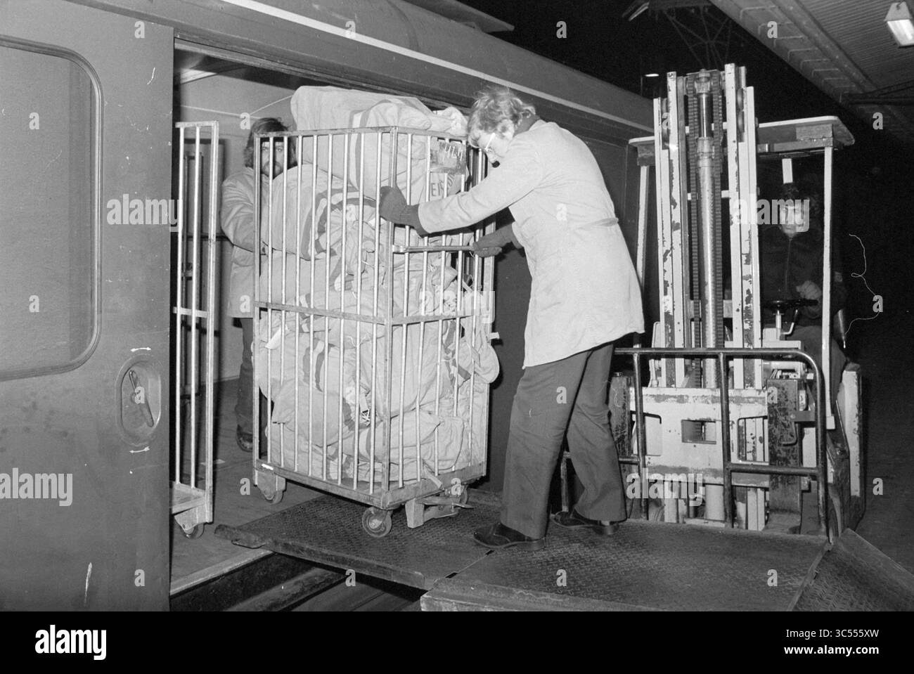 Sortieren von Post, Post Baljuwslaan, PTT, Post, Stadtpost, Haarlem, Baljuwslaan, Niederlande, 21-12-1978 Whizgle News, Dutch Desk, Niederlande, 1950-2000 Ein Arbeiter entlädt vorsichtig einen großen Wagen mit gebündeltem Material aus einem Zug, während ein Gabelstaplerfahrer in der Nähe wartet, um beim Ladevorgang zu helfen. Die Szene findet bei Nacht statt, beleuchtet durch Stationslichter. Stockfoto