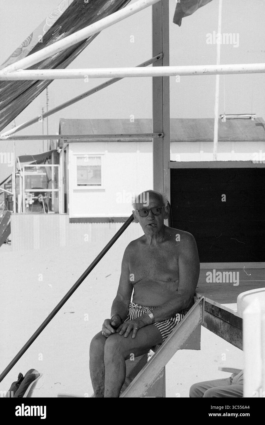 Onkel Joop, Talk of the Day, 10-07-1991 Whizgle News, Dutch Desk, Niederlande, 1950-2000 ein älterer Mann sitzt auf einer hölzernen Plattform, trägt Badehosen und Brillen und blickt nachdenklich in die Ferne. Im Hintergrund steht ein Strandhaus, umgeben von einer sandigen Landschaft. Stockfoto