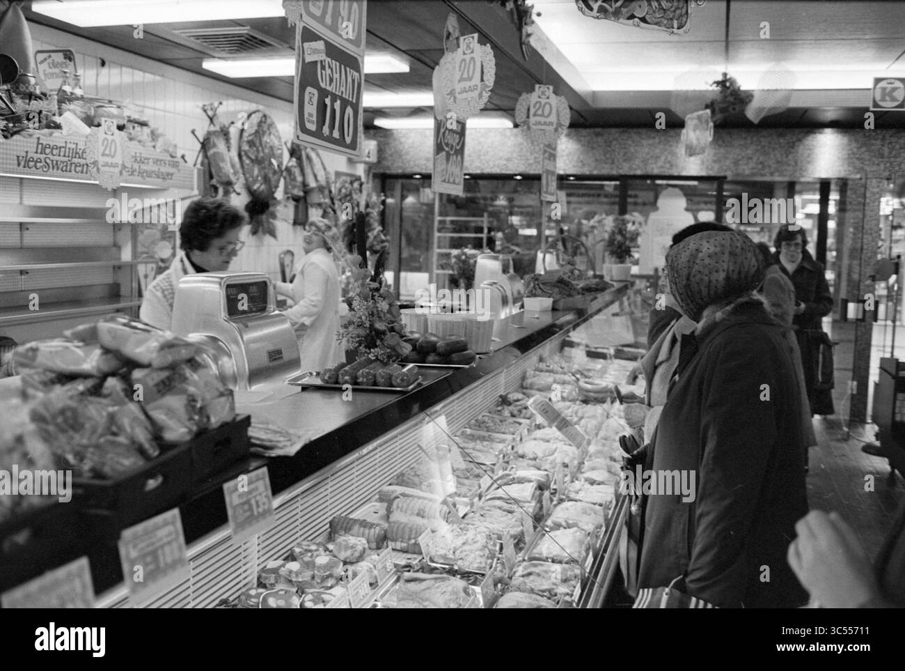 Geschäftsbericht Slager v.d. Geest (Interior Store), 01-12-1980 Whizgle News, Dutch Desk, Niederlande, 1950-2000 Eine geschäftige Marktszene mit einer Theke voller verpackter Waren und frischer Artikel. Käufer durchsuchen die Auswahl eifrig, während ein Mitarbeiter mit Kunden interagiert. Farbenfrohe Schilder zeigen Preise an, was zur lebhaften Atmosphäre des Marktplatzes beiträgt. Stockfoto
