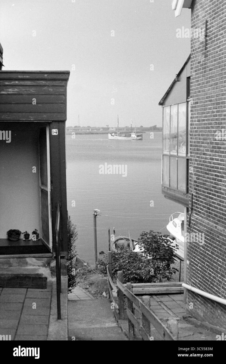 Ansicht des IJ von Spaarndam, Spaarndam, 20-08-1979 Whizgle News, Dutch Desk, Niederlande, 1950-2000 Eine ruhige Szene mit einem schmalen Pfad, der zwischen zwei Gebäuden nach unten führt, mit Blick auf ein ruhiges Gewässer in der Ferne. Ein Boot liegt vor Anker, während die nahe gelegenen Laubbäume und Stufen zur friedlichen Atmosphäre beitragen. Stockfoto