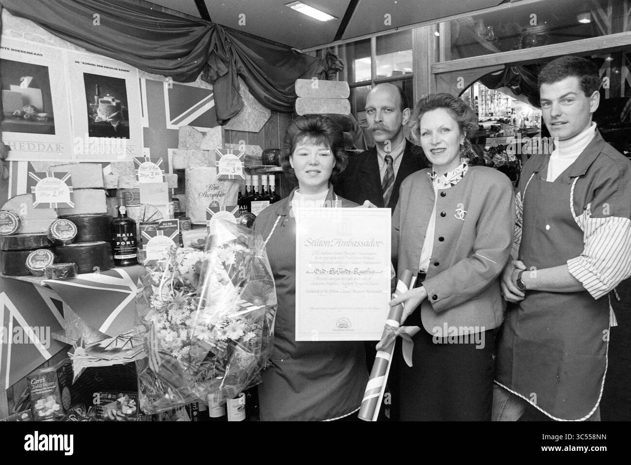 Preis altes niederländisches Käsehaus, Schalkwijk, Haarlem, Niederlande, 20-02-1992 Whizgle News, Dutch Desk, Niederlande, 1950-2000 Eine Gruppe von vier Personen steht stolz in einem Geschäft und stellt ein großes Zertifikat und einen Blumenstrauß vor. Hinter ihnen befinden sich Regale mit einer Auswahl an Käse und Weinen, umgeben von Dekorationen, die die britische Kultur zelebrieren. Die Atmosphäre vermittelt ein Gefühl der Leistung und des Feierns. Stockfoto