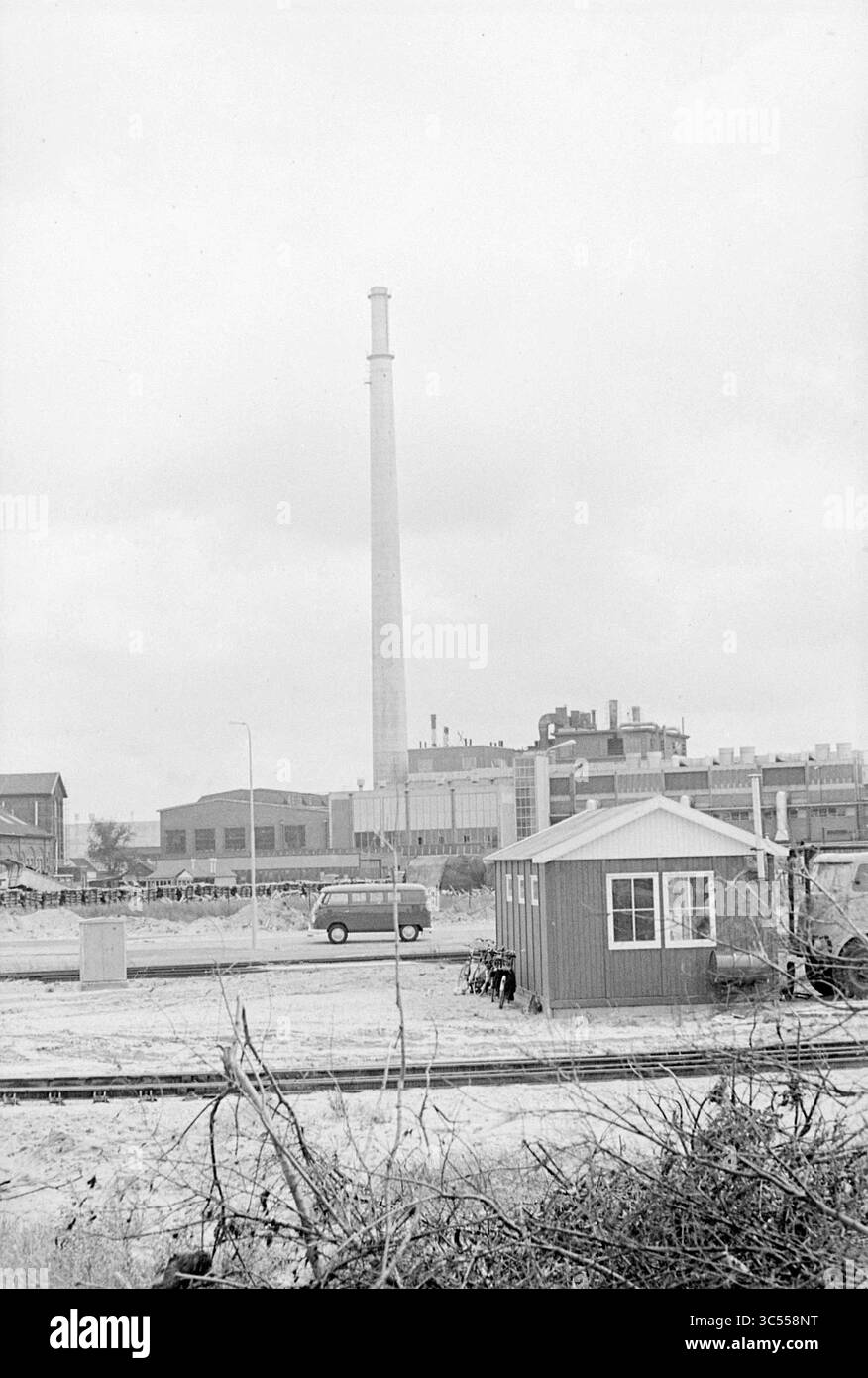 Chimney P Velzen van Gelder Hoogovens, Buildings, 12-08-1963 Whizgle News, Dutch Desk, Niederlande, 1950-2000 Ein hoher Schornstein erhebt sich über einer Industrielandschaft, umgeben von Gebäuden und einem kleinen Schuppen. Die Bahngleise führen durch den Vordergrund, was auf die funktionierende industrielle Tätigkeit des Gebiets hindeutet. In der Ferne bilden verschiedene Strukturen eine Kulisse vor einem bewölkten Himmel. Stockfoto