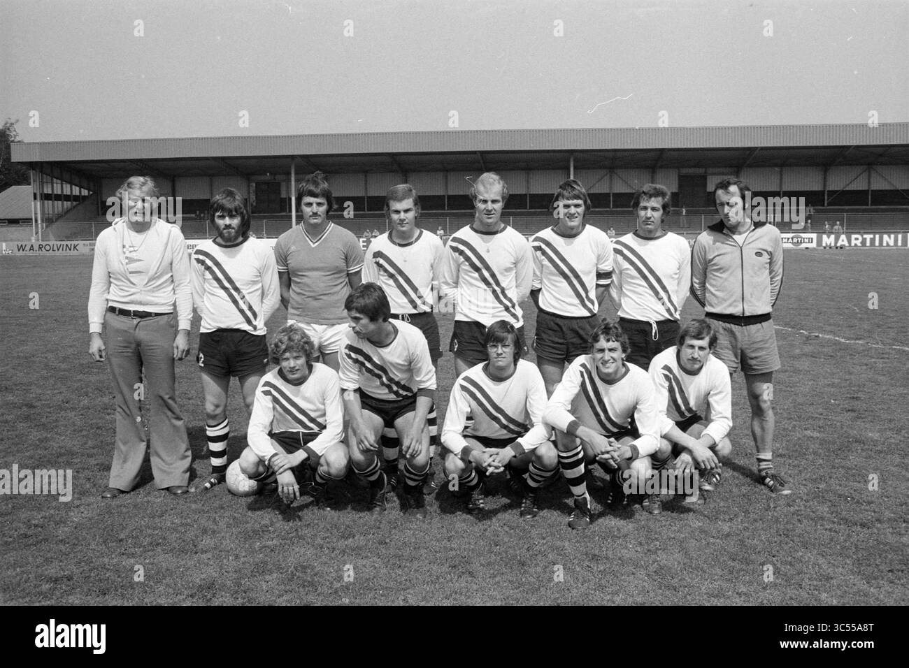 IJmuiden - Vroomshoopse Boys, Football IJmuiden, 05-06-1976 Whizgle News, Dutch Desk, Niederlande, 1950-2000 Eine Fußballmannschaft posiert auf dem Spielfeld in passenden Uniformen mit auffälligen Diagonalstreifen. Sie stehen in zwei Reihen, mit dem Coach oder Manager auf der linken Seite. Der Hintergrund zeigt eine Stadionstruktur und Werbetafeln, die auf einen Wettbewerbsgeist hinweisen. Stockfoto