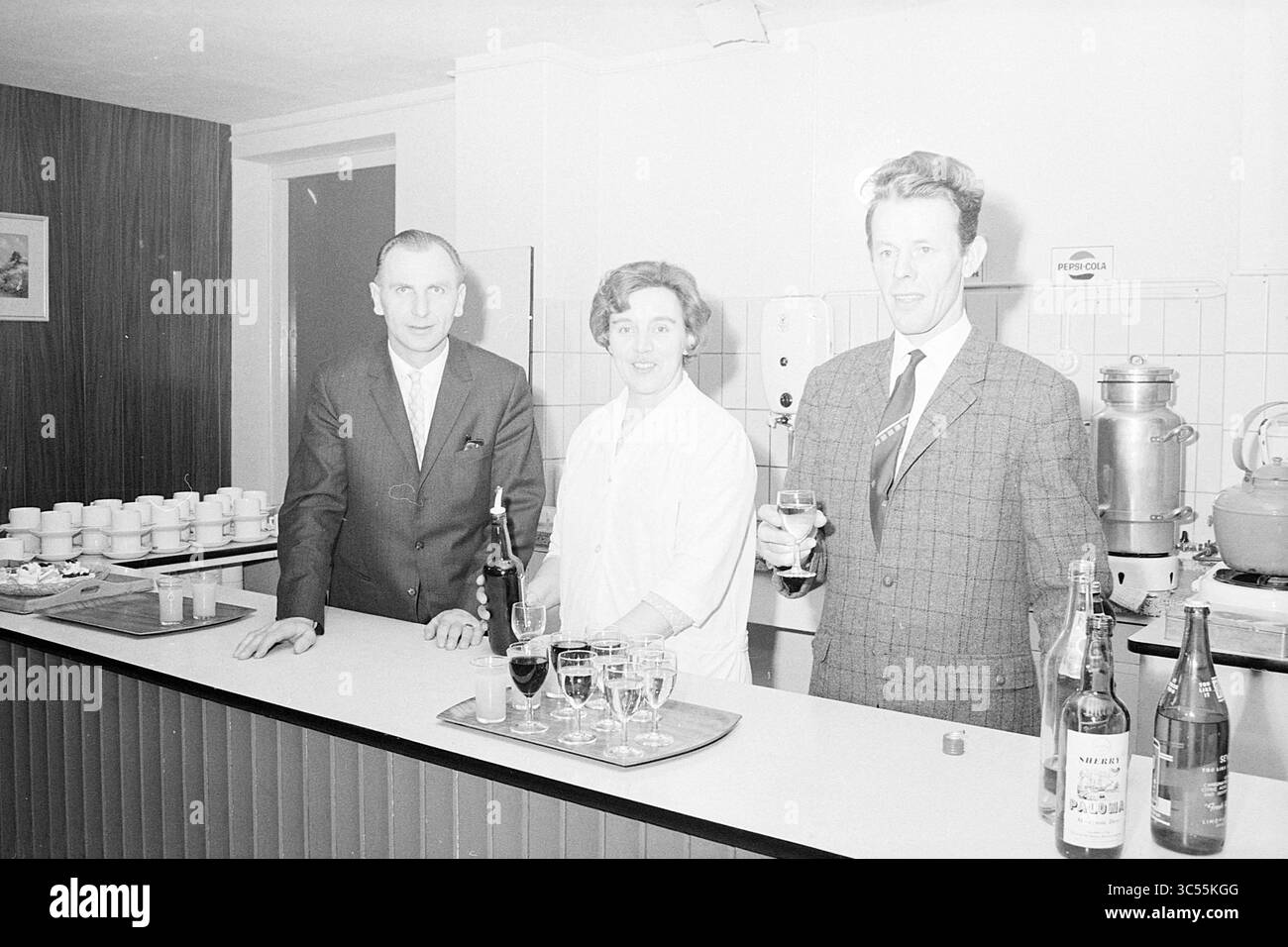 Jopie the Barmaid, Blast Furnace Chess Tournament, 28-01-1969 Whizgle News, Dutch Desk, Niederlande, 1950-2000 drei Personen stehen hinter einem Bartheken, jeder hält ein Glas. Der Mann links trägt einen Anzug und lächelt, während die Frau in der Mitte fröhlich in einem weißen Outfit wirkt. Der Mann auf der rechten Seite trägt einen karierten Blazer und eine Krawatte. Verschiedene Flaschen sind auf dem Tresen zu sehen, was zu einer festlichen Atmosphäre beiträgt. Hinter ihnen ist eine Wand mit Bechern und anderen Gegenständen geschmückt, was auf ein geselliges Treffen hindeutet. Stockfoto