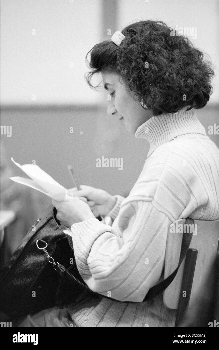 Stadtrat H. Elmar, Green Left, 01-11-1993 Whizgle News, Dutch Desk, Niederlande, 1950-2000 Eine junge Frau mit lockigem Haar sitzt und konzentriert sich auf ein Blatt Papier in ihren Händen. Sie trägt einen kuscheligen Rollkragenpullover und hat einen Bleistift, was darauf hindeutet, dass sie sich entweder Notizen macht oder an etwas Wichtigem arbeitet. Eine Handtasche liegt an ihrer Seite und trägt zu einer fleißigen Atmosphäre bei. Stockfoto