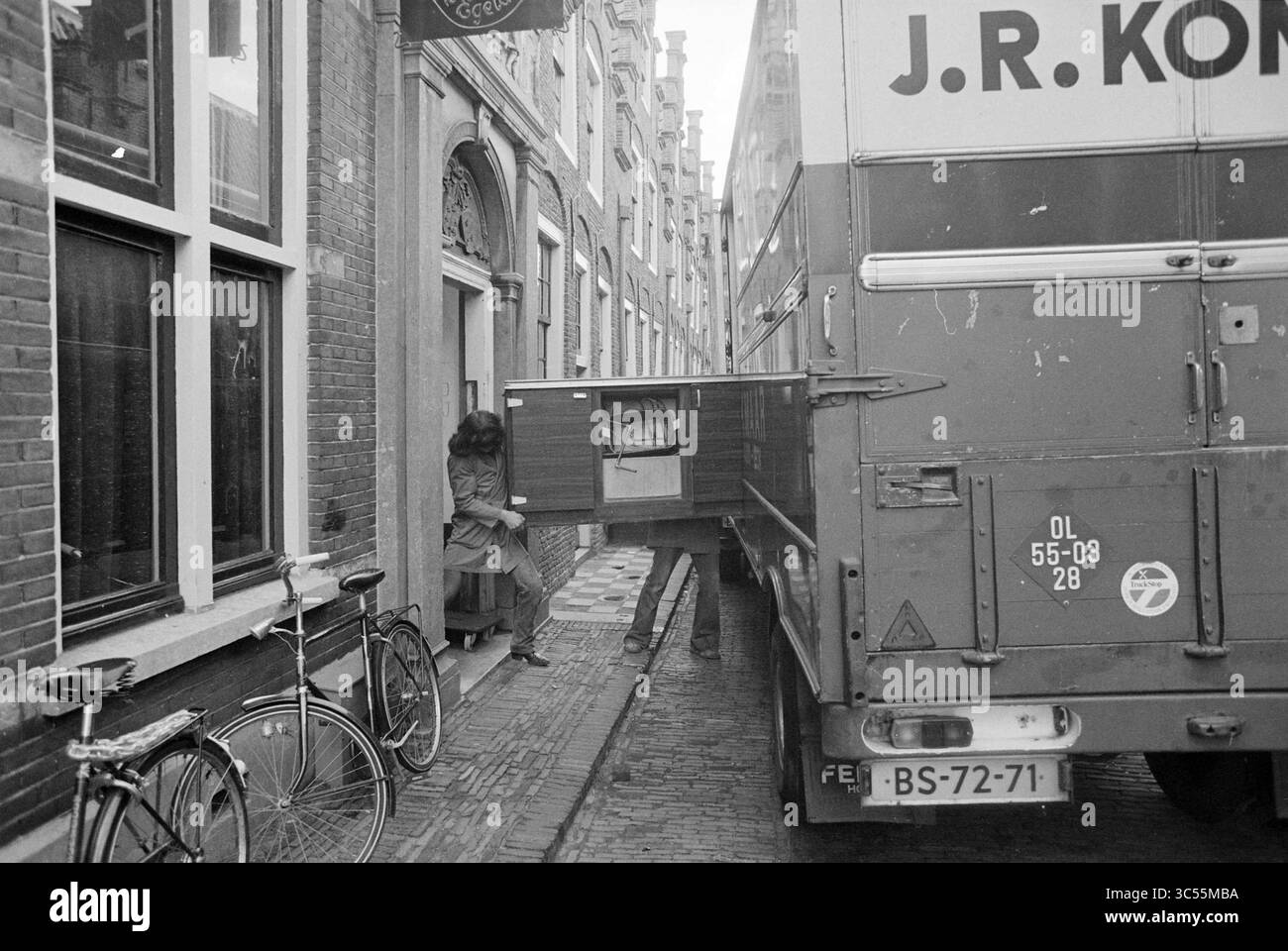 Clearing Café, Kulturzentrum de Egelantier (fahrender Van auf der Straße), Haarlem, Groot Heiligland, Niederlande, 20-11-1978 Whizgle News, Dutch Desk, Niederlande, 1950-2000 Eine Frau navigiert vorsichtig durch die enge Straße, während ein fahrender Lastwagen ein Möbelstück mit einem in der Nähe geparkten Fahrrad und historischen Gebäuden entlang der Straße lädt. Stockfoto