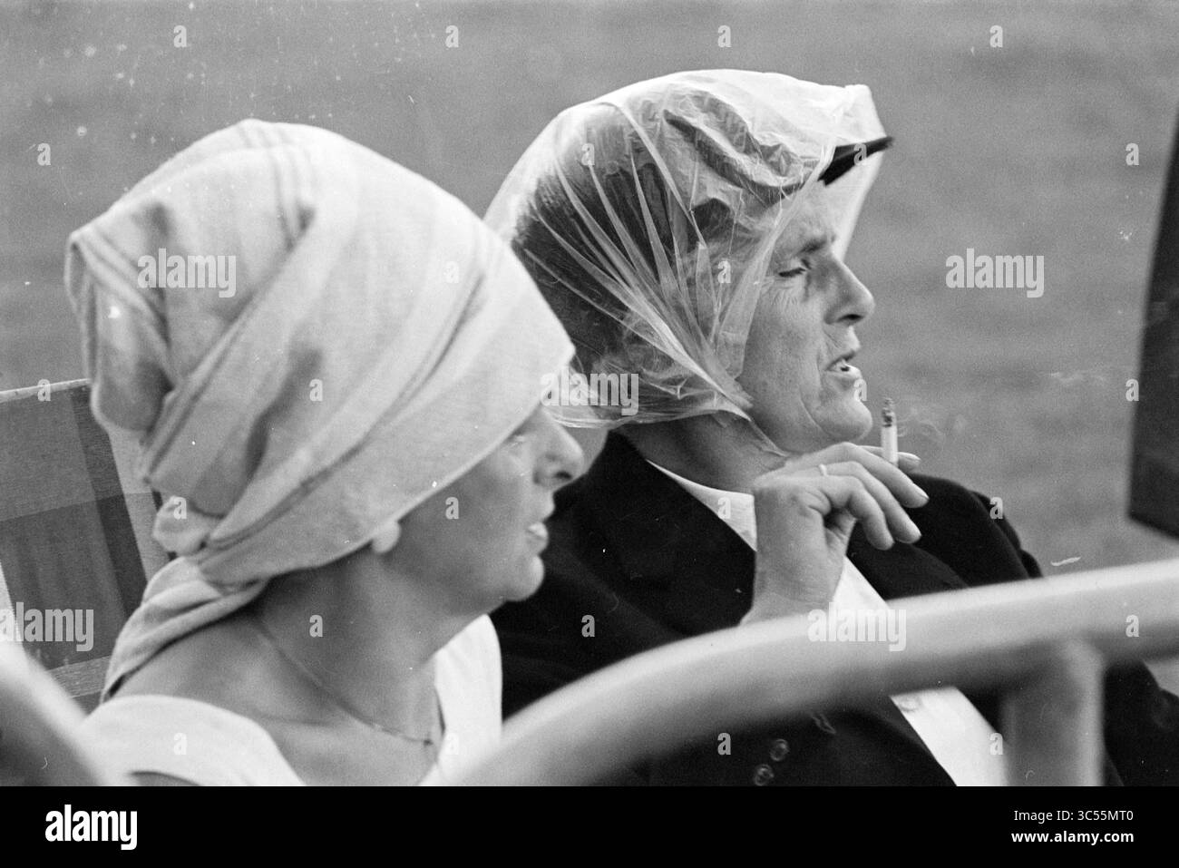 Reitwettbewerb, Traberpferde, 08-07-1970 Whizgle News, Dutch Desk, Niederlande, 1950-2000 zwei Personen sitzen nebeneinander, beide tragen Kopfbedeckung. Eine Person, die leicht abgewandt ist, hat ihr Gesicht meist verdeckt, während die andere, ein älteres Individuum, eine Zigarette raucht und tief in Gedanken erscheint. Die Umgebung lässt eine ungezwungene Outdoor-Umgebung vermuten, und die Stimmung wirkt reflektierend. Stockfoto