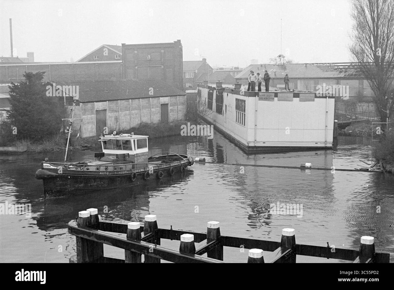 Das alte ausgebrannte Gemeindezentrum (altes Schulboot) blockierte den Schiffsverkehr auf der Spaarne bei der Prinsenbrug; aus Protest gegen die Gemeinde Haarlem schleppten die Bewohner der Harmenjanswijk das Boot auf die Spaarne, um ihren Wunsch nach einem neuen Gemeindezentrum zu bekräftigen., Haarlem, Niederlande, 23-03-1980 Whizgle News, Dutch Desk, Niederlande, 1950-2000 Ein kleines Boot fährt durch ruhiges Wasser in der Nähe eines teilweise errichteten Gebäudes, wo mehrere Personen auf dem Dach stehen, während andere vom Rand des Gebäudes aus beobachten. Umliegende Lagerhäuser und Industriegebäude entstehen Stockfoto