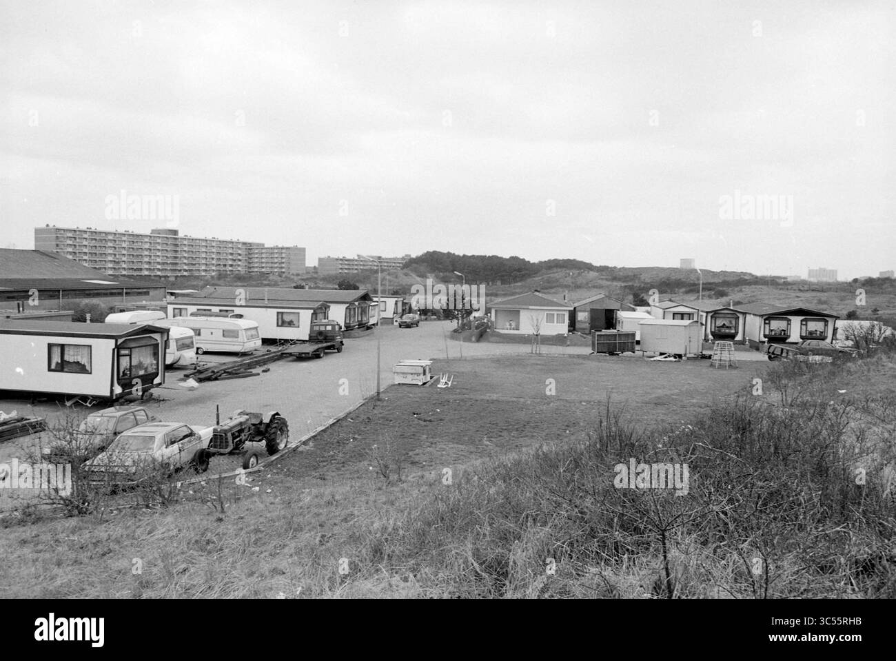 Trailer Camp, Zandvoort, Zandvoort, 05-04-1993 Whizgle News, Dutch Desk, Niederlande, 1950-2000 Eine Gruppe von Anhängern und provisorischen Bauwerken ist in einem offenen Gebiet angeordnet, umgeben von karger Vegetation und niedrigen Hügeln. Im Hintergrund erhebt sich eine Reihe von Wohngebäuden vor einem bewölkten Himmel und trägt zu einem Gefühl eines Übergangsraums bei. Stockfoto