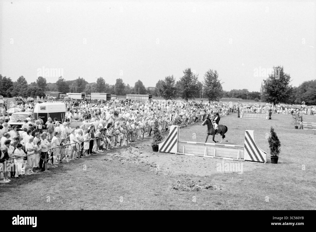 Reitwettbewerb Whizgle News, Dutch Desk, Niederlande, 1950-2000 Eine große Menschenmenge versammelt sich, um einen Reiter zu Pferd zu beobachten, wie er in einer Reitveranstaltung, eingerahmt von Bäumen und offenen Feldern, durch ein Hindernis navigiert. Stockfoto
