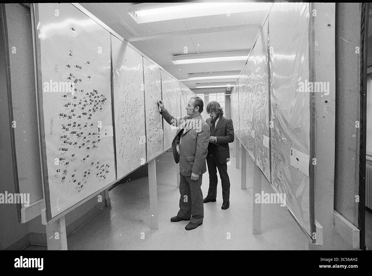 Int. North Holland Sports Council, Interior, 22.01-1974 Whizgle News, Dutch Desk, Niederlande, 1950-2000 zwei Personen analysieren in einem geräumigen Raum detaillierte Karten, die an den Wänden angebracht sind. Eine Person zeigt auf einen bestimmten Bereich im Diagramm, während die andere genau beobachtet. Die Umwelt schlägt ein akademisches oder Forschungsumfeld vor, wobei der Schwerpunkt auf Datenpräsentation und -Analyse liegt. Stockfoto