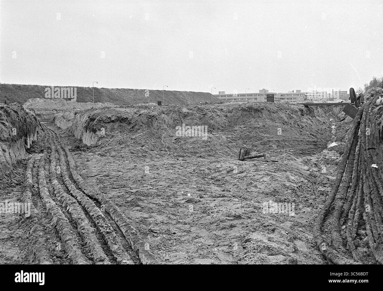 GroundWorks Beethovenstraat, Boelelaan in Amsterdam., de Ruiter B.V. Halfway, Amsterdam, Beethovenstraat, Niederlande, 23-10-1974 Whizgle News, Dutch Desk, Niederlande, 1950-2000 Ein großer, mit schlammigem Boden gefüllter Aushub mit tiefen Reifenspuren und Maschinen im Hintergrund. Die Landschaft wird von Erdbauarbeiten dominiert, was auf Bau- oder Landschaftsgestaltung in der Nähe hindeutet. In der Ferne sind Gebäude vor bewölktem Himmel zu sehen. Stockfoto