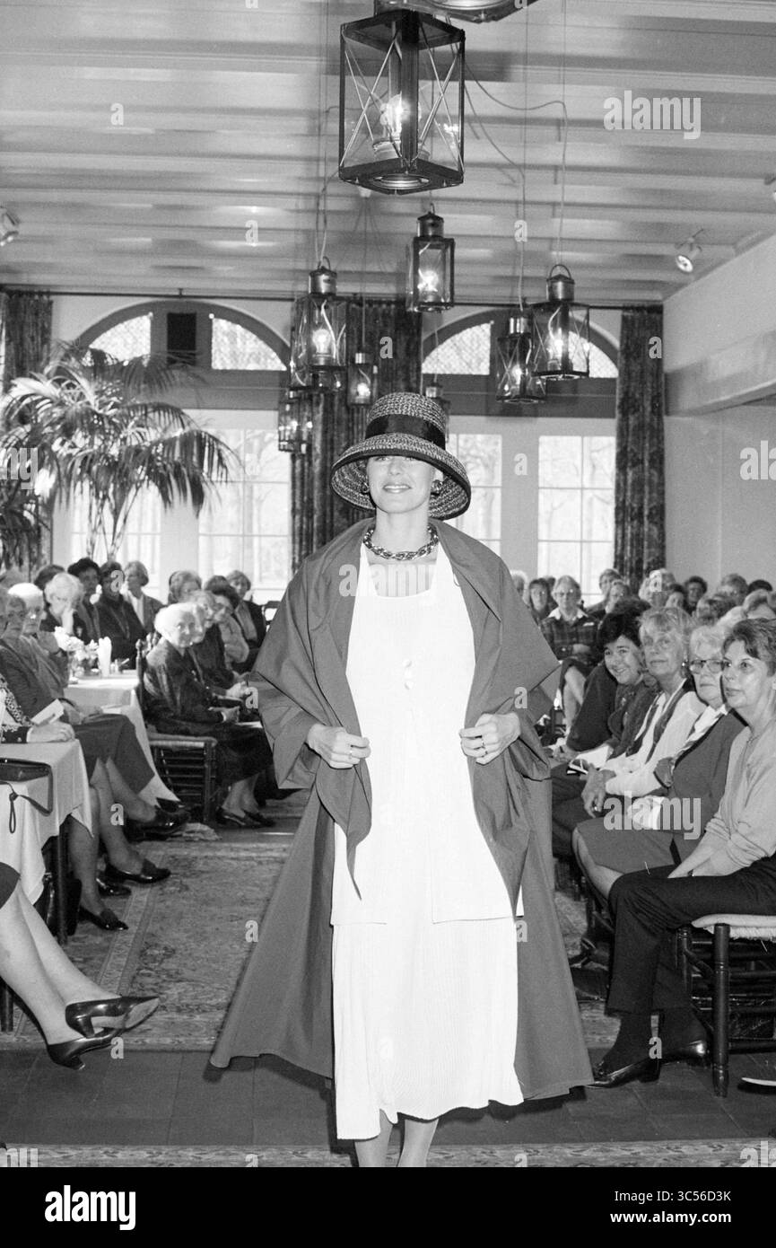 Fashion Show, 00-00-1995 Whizgle News, Dutch Desk, Niederlande, 1950-2000 Ein selbstbewusstes Modell zeigt ein elegantes Outfit, ergänzt durch einen stilvollen Hut und ein fließendes cape, während ein Publikum von Frauen aufmerksam in einem eleganten Ambiente mit hängenden Laternen und üppigem Grün beobachtet. Stockfoto