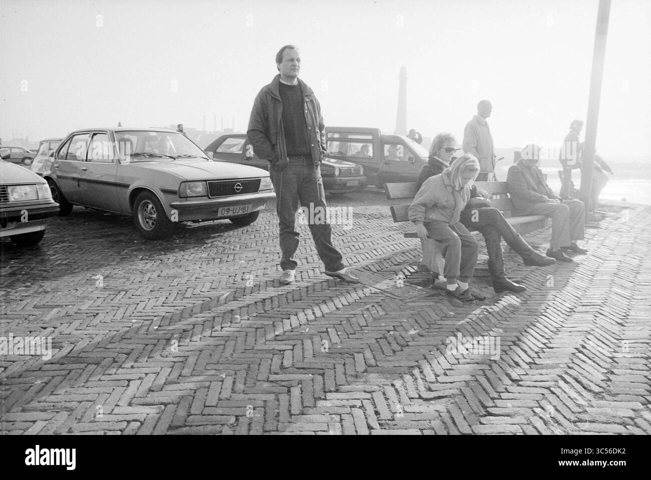Geschäftiger Leiter des Hafens, Ports, 08-02-1989 Whizgle News, Dutch Desk, Niederlande, 1950-2000 Eine Gruppe von Menschen sitzt beiläufig auf einer Bank, während ein Mann in der Nähe steht und die Szene um sie herum betrachtet. Im Hintergrund parken Oldtimer auf einem Kopfsteinpflasterweg, was auf eine ruhige Atmosphäre hindeutet. Die entfernte Silhouette einer Struktur ist sichtbar und verleiht der Einstellung ein Gefühl der Tiefe. Stockfoto