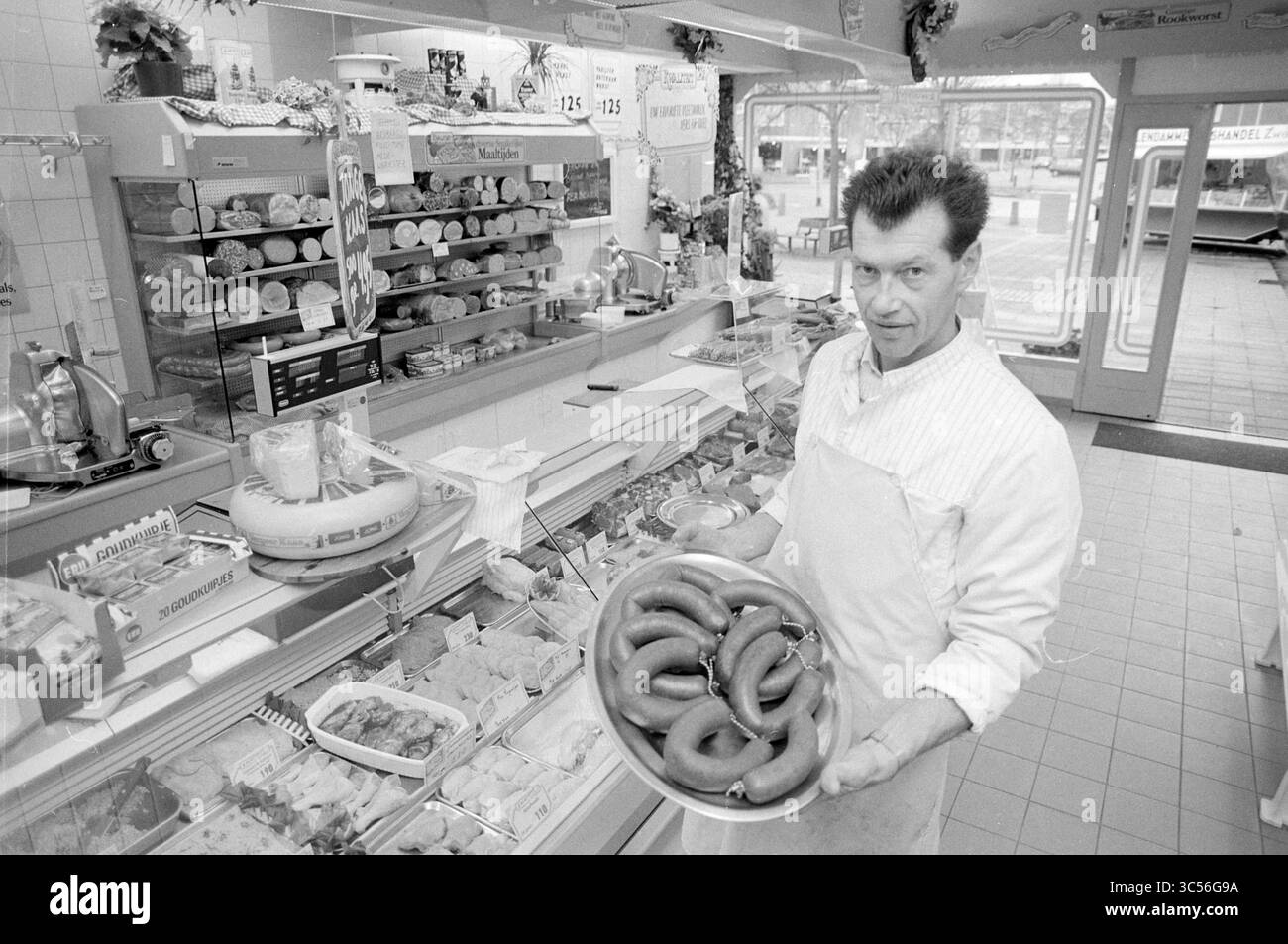 Metzgerei des Landes, H'lem, Haarlem, Niederlande, 07-11-1991 Whizgle News, Dutch Desk, Niederlande, 1950-2000 Ein Ladenbesitzer steht stolz hinter einer Auswahl an Fleisch- und Köstlichkeiten und hält ein Tablett mit Würstchen. Das geschäftige Geschäft bietet eine Auswahl an Produkten, mit Regalen mit verschiedenen verpackten Waren und frischen Artikeln, die eine lebhafte Atmosphäre schaffen. Stockfoto