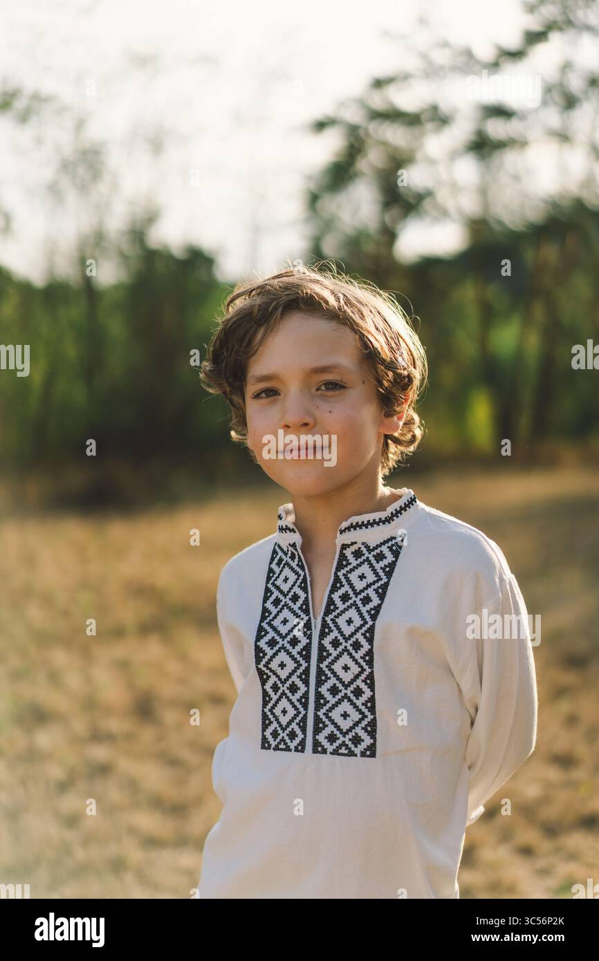 Ein Junge lächelt selbstbewusst, während er auf einem sonnendurchfluteten Feld steht und ein traditionelles besticktes Hemd trägt Stockfoto