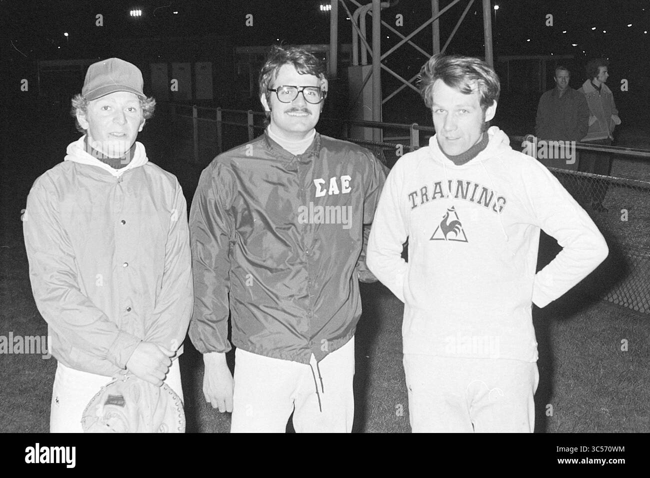 Amerikanische Baseballspieler Hit Nicols, Baseball, 12-04-1973 Whizgle News, Dutch Desk, Niederlande, 1950-2000 drei Männer posieren in der Nacht zusammen im Freien und stehen Seite an Seite mit lässiger Kleidung. Einer trägt eine Mütze und eine Jacke, während ein anderer einen Schnurrbart und eine Jacke mit den Buchstaben „CAE“ trägt. Der dritte Mann trägt ein Sweatshirt mit dem Wort „TRAINING“. Ein schwaches Licht aus der Ferne erzeugt eine Kulisse, die auf eine lebhafte Umgebung in der Nähe hindeutet. Stockfoto