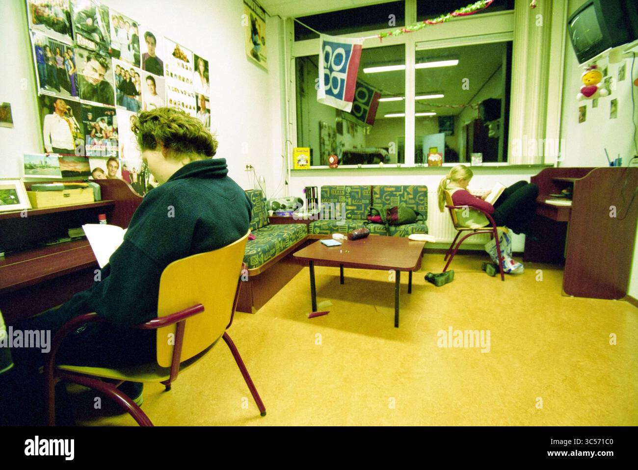 Internatsstudienzeit, IJmuiden, IJmuiden, Niederlande, 21.01.2002 Whizgle News, Dutch Desk, Niederlande, 1950-2000 zwei Personen nehmen ihre eigenen Aktivitäten in einem gemütlichen Gemeinschaftsraum wahr. Eine Person sitzt an einem Schreibtisch und liest, während die andere sich auf einem Stuhl in der Nähe entspannt. Die Wände sind mit verschiedenen Fotografien und Dekorationen verziert, was zu einer einladenden Atmosphäre beiträgt. Eine Couch und ein Couchtisch runden das Ambiente ab und betonen eine Mischung aus persönlichen und gemeinsamen Momenten. Stockfoto