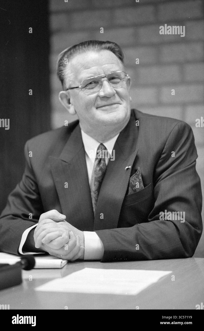 New Council Alberts, Bennebroek, Bennebroek, 25-03-1992 Whizgle News, Dutch Desk, Niederlande, 1950-2000 Ein gut gekleideter Mann mit Brille sitzt selbstbewusst an einem Tisch, lächelt leicht, während er seine Hände aneinander legt. Er trägt Anzug und Krawatte, begleitet von einem gemusterten Taschenfeld, was auf einen formellen Anlass oder ein wichtiges Treffen hindeutet. Der Hintergrund ist mit einer Ziegelwand versehen, die der Umgebung einen Hauch von Textur verleiht. Stockfoto