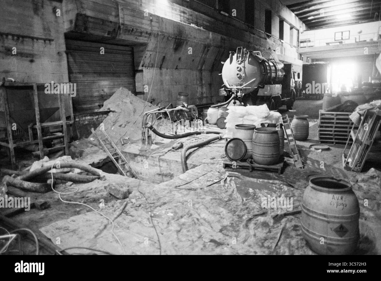 Arbeit am IJ Bunker und unterirdischen Keller mit Kontamination, Bunker, IJmuiden, Nederland, 06-12-1988 Whizgle News, Dutch Desk, Niederlande, 1950-2000 ein Industrieraum mit Ausrüstung und Fässern, mit einem großen Tankwagen und einem Bauort oder einer Renovierung. Werkzeuge und Materialien sind verstreut, was auf eine aktive Arbeitsumgebung hindeutet. Licht strömt von einer Tür herein und beleuchtet den Arbeitsbereich. Stockfoto