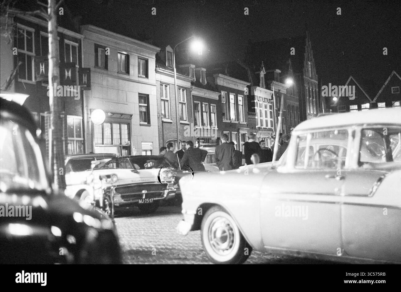Straßenszene. Car Trade, Haarlem, Botermarkt, Niederlande, 07-11-1963 Whizgle News, Dutch Desk, Niederlande, 1950-2000 Eine geschäftige nächtliche Szene mit klassischen Autos, die entlang einer kopfsteingepflasterten Straße geparkt werden, mit alten Gebäuden im Hintergrund. Gruppen von Personen unterhalten sich, beleuchtet von Straßenlaternen, die die lebhafte Atmosphäre in warmen Farben überstrahlen. Stockfoto