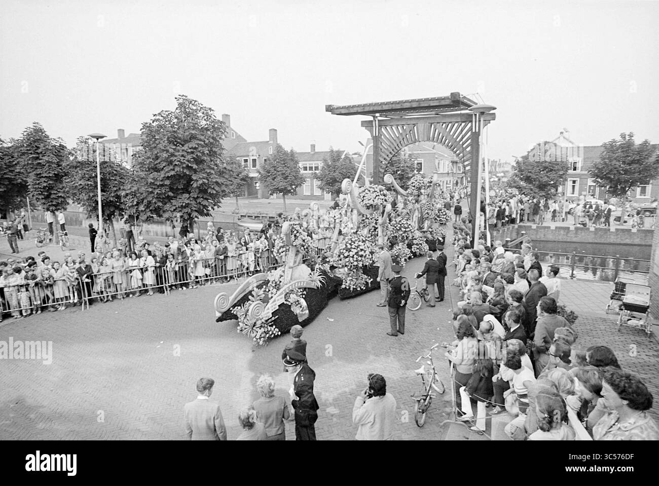 Blumenparade Aalsmeer, Blumenparade, Blumenausstellung, 04-09-1971 Whizgle News, Dutch Desk, Niederlande, 1950-2000 Eine lebhafte Paradeszene mit einem reich verzierten Wagen mit Blumenschmuck entfaltet sich, während sich hinter Barrieren eine Menschenmenge versammelt, die die Festlichkeiten sehnt. Bäume säumen die Straße, und ein großes Bauwerk im Hintergrund umrahmt die Prozession und verleiht der Veranstaltung eine festliche Atmosphäre. Menschen jeden Alters, einige auf Fahrrädern, sind an der Feier beteiligt. Stockfoto