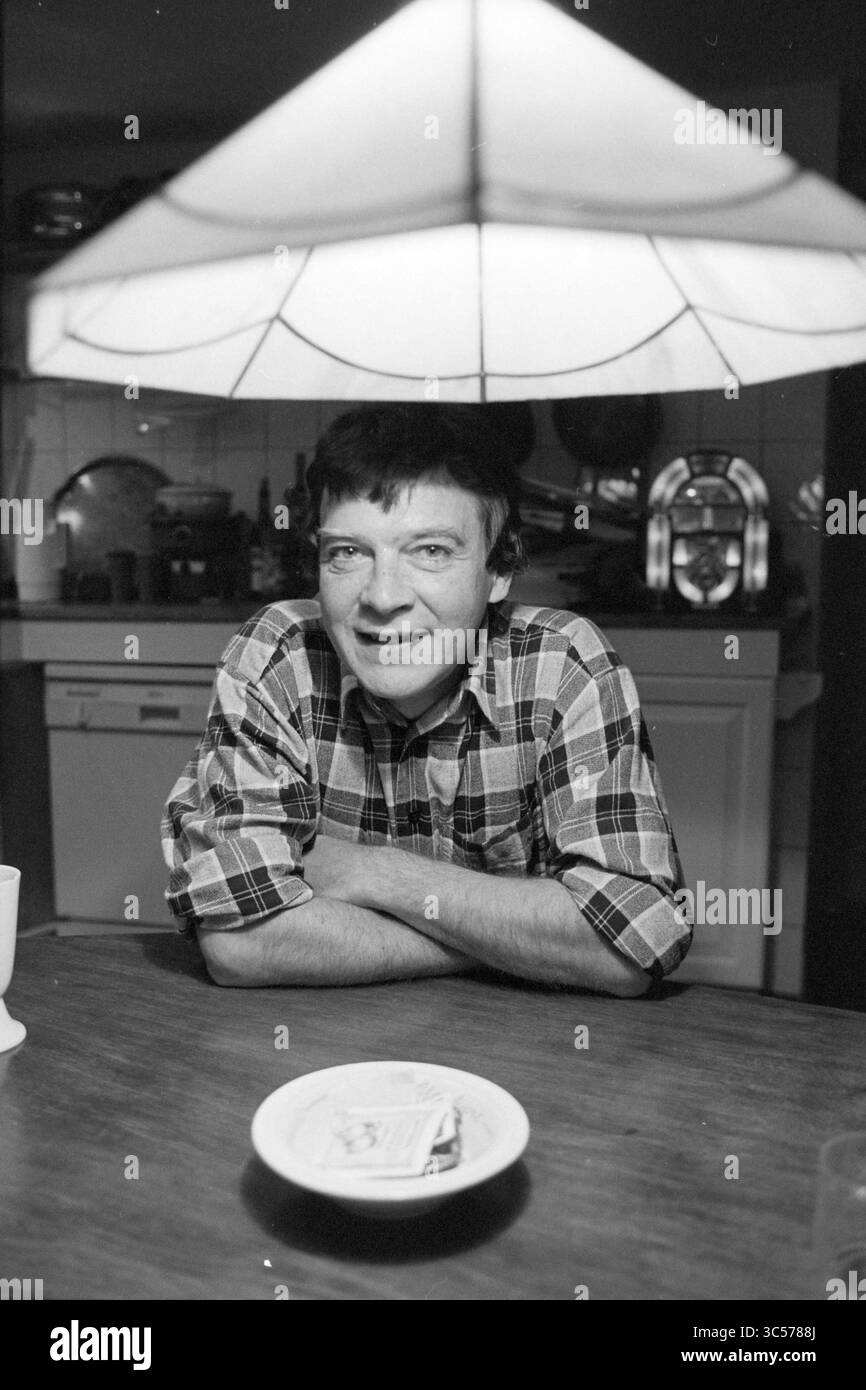 Koos Pelsser, Green Left, 30-10-1996, Whizgle News, Dutch Desk, Niederlande, 1950-2000 Ein Mann mit einem freundlichen Lächeln sitzt an einem Tisch, beleuchtet von einer Hängelampe. Er trägt ein kariertes Hemd und legt seine Arme auf den Tisch, der mit einem kleinen Teller versehen ist. Im Hintergrund schaffen Küchenutensilien eine gemütliche Atmosphäre. Stockfoto