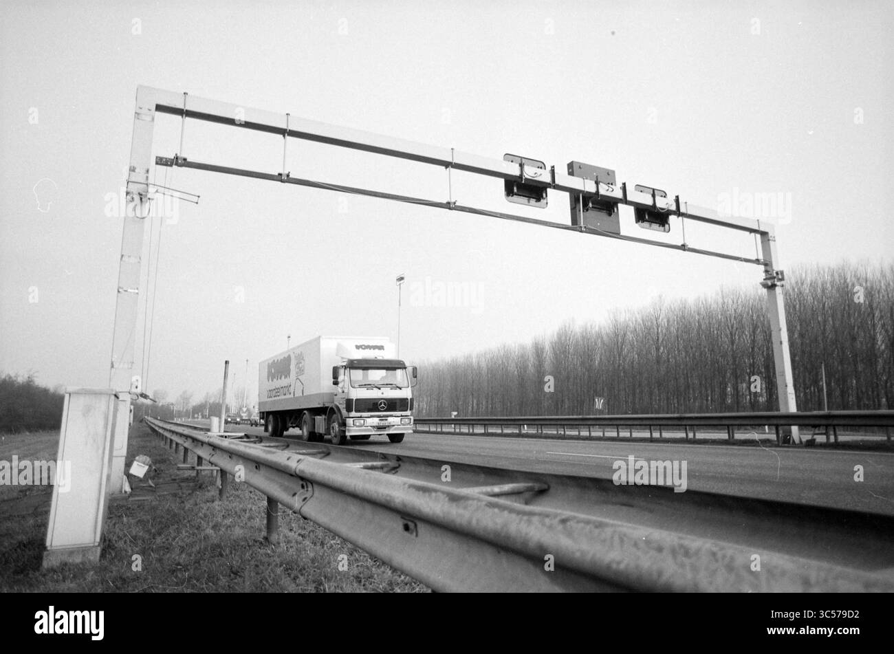 Höhenmesser am Velsertunnel, 05-02-1992 Whizgle News, Dutch Desk, Niederlande, 1950-2000 Ein großer Lkw fährt auf einer Autobahn unter einer mit Verkehrssignalen oder Kameras ausgestatteten Dachkonstruktion vorbei. Spärliche Bäume säumen die Straße und schaffen eine ruhige, offene Atmosphäre. Stockfoto