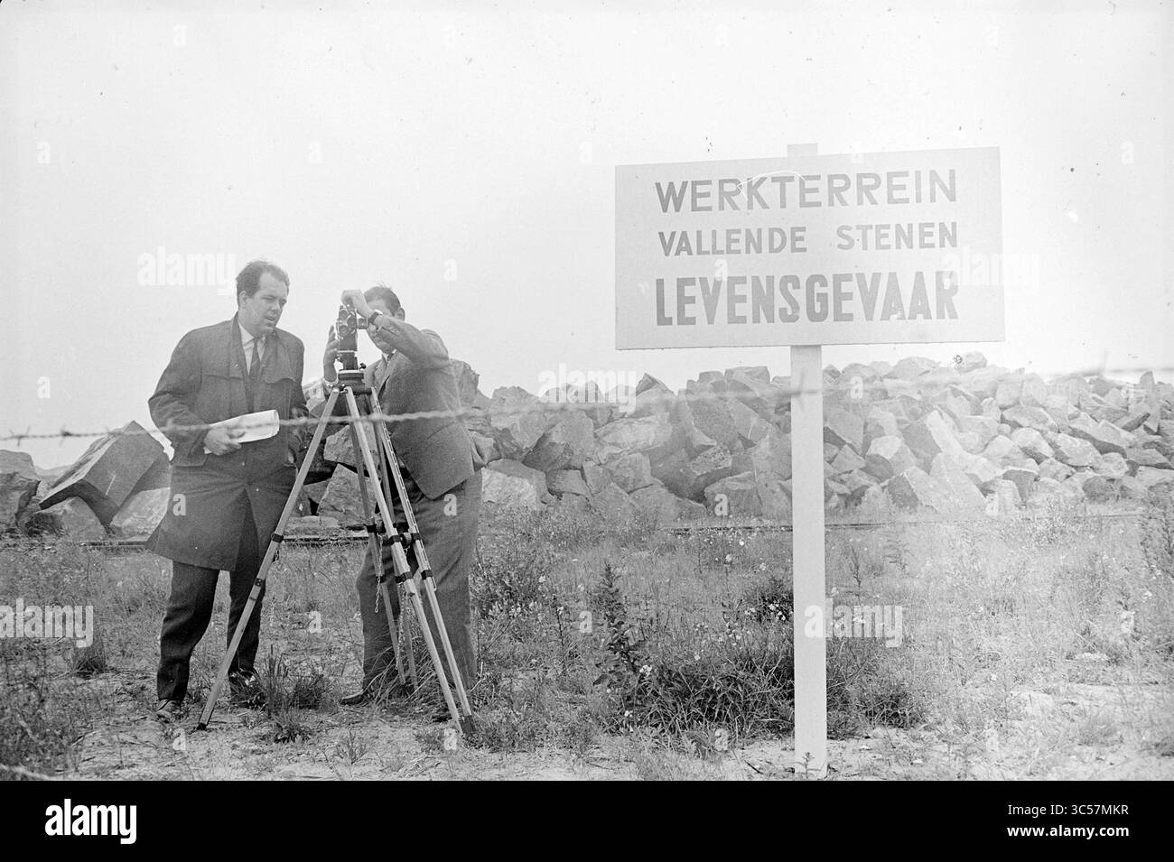 Piet van Strien und Wilco Nehus Film über Piers, Dreharbeiten, Filmaufnahmen, 06-07-1962 Whizgle News, Dutch Desk, Niederlande, 1950-2000 zwei Männer untersuchen ein Vermessungsinstrument in der Nähe einer Baustelle, mit einem Schild, das vor herabfallenden Steinen warnt. Gesteine und Schutt sind überall verstreut, was auf laufende Arbeiten und potenzielle Gefahren hinweist. Stockfoto