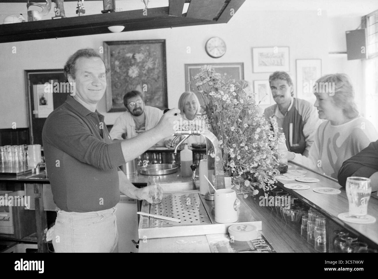 Paul van Egmond in Café, Café, Haarlem, kleine Houtstraat, Niederlande, 02-07-1985 Whizgle News, Dutch Desk, Niederlande, 1950-2000 Eine lebhafte Bar-Szene fängt einen lächelnden Barkeeper ein, der Getränke einschenkt, umgeben von begeisterten Gästen am Theken. Blumenarrangements und zwanglose Gespräche verstärken die warme Atmosphäre der Kameradschaft und des Genusses im Hotel. Stockfoto