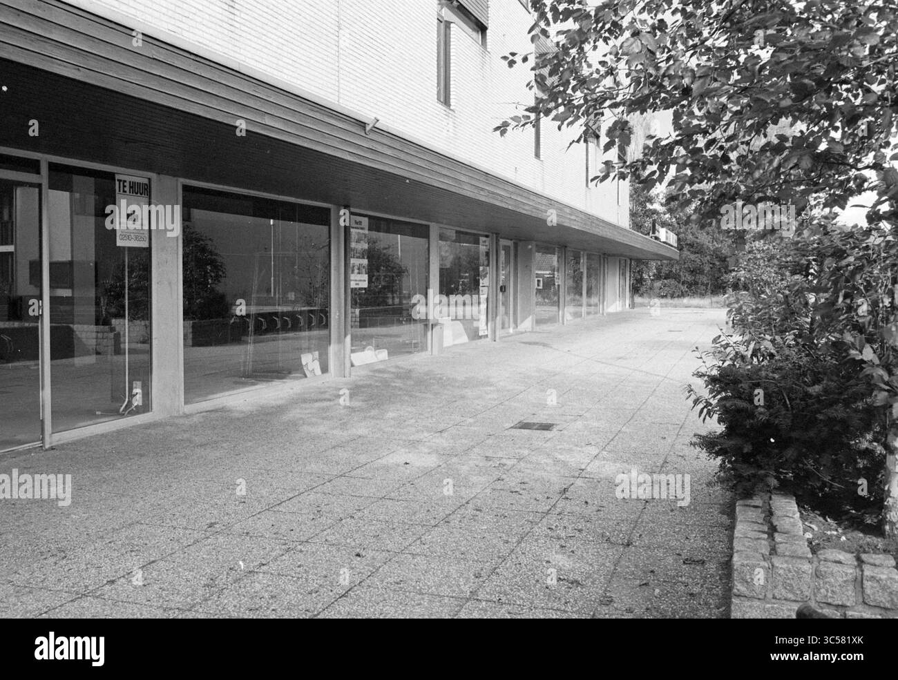 Leere Geschäfte Nielenplein Heemskerk, Heemskerk, Geschäfte und Kioske, Heemskerk, Burgemeester Nielenplein, 14-10-1981 Whizgle News, Dutch Desk, Niederlande, 1950-2000 unter einem modernen Überhang befindet sich Ein leeres Schaufenster mit großen Glasfenstern, umgeben von einem gepflasterten Fußweg und Grün. Die Gegend scheint ruhig zu sein, und in der leeren Fläche sind Anzeichen von früherer Aktivität zu erkennen. Stockfoto