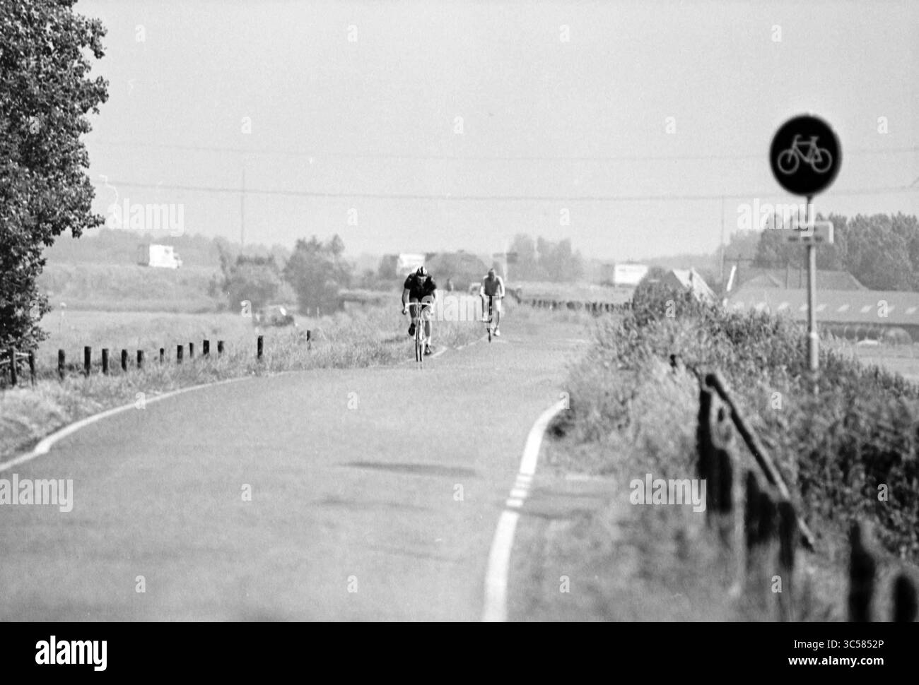 Cyclists, Sp'woude, Spaarnwoude, 13-06-1994 Whizgle News, Dutch Desk, Niederlande, 1950-2000 zwei Radfahrer fahren entlang einer gewundenen Straße, flankiert von Bäumen und einem niedrigen Zaun, mit weit entfernten Feldern und Gebäuden im Hintergrund. In der Nähe befindet sich ein Schild für den Fahrradweg, das auf die gemeinsame Nutzung der Straße hinweist. Stockfoto