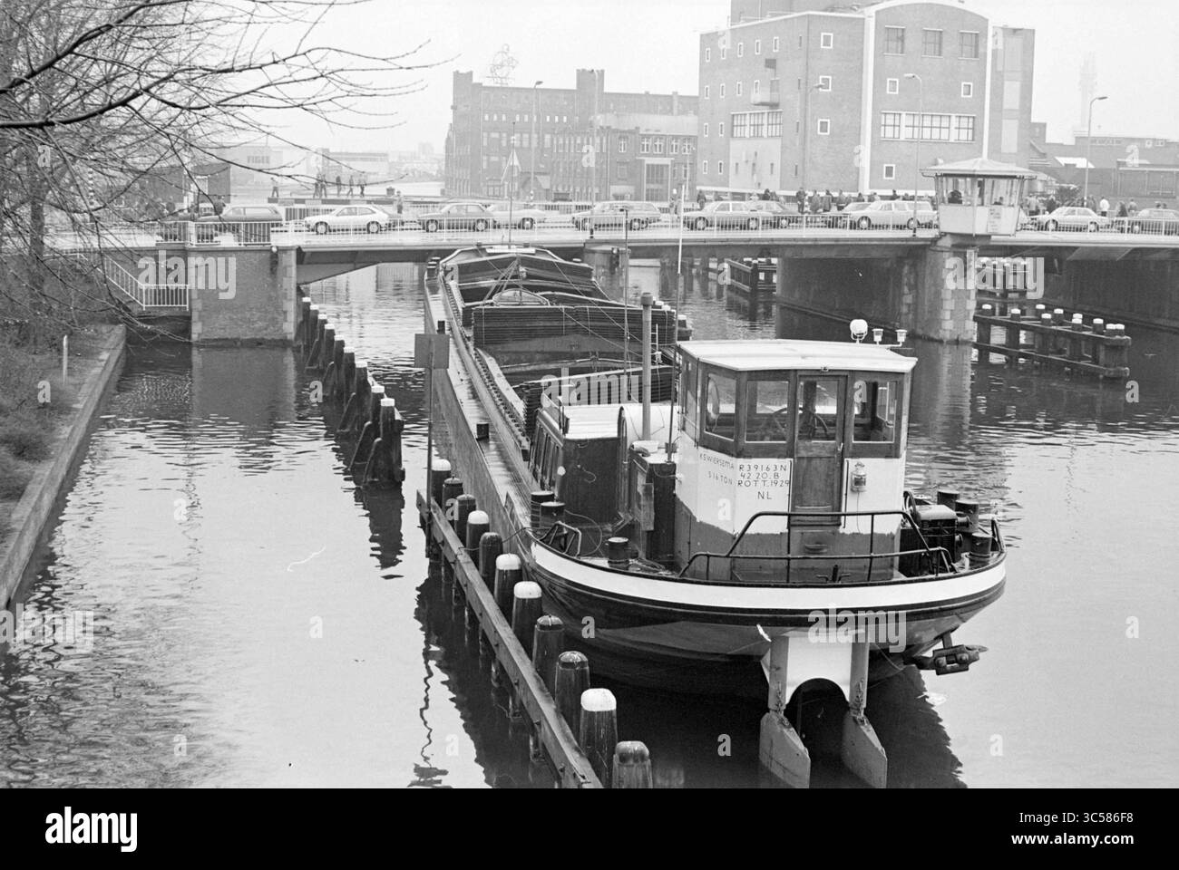 Das alte ausgebrannte Gemeindezentrum (altes Schulboot) blockierte den Schiffsverkehr auf der Spaarne bei der Prinsenbrug; aus Protest gegen die Gemeinde Haarlem schleppten die Bewohner der Harmenjanswijk das Boot auf die Spaarne, um ihren Wunsch nach einem neuen Gemeindezentrum zu bekräftigen., Haarlem, Niederlande, 23-03-1980 Whizgle News, Dutch Desk, Niederlande, 1950-2000 Ein Boot liegt an einem Dock, dessen Kanal sanft die umgebenden Strukturen reflektiert. Im Hintergrund überspannt eine Brücke die Wasserstraße, während mehrere Autos in der Nähe parken, was auf eine geschäftige städtische Umgebung hindeutet. Stockfoto