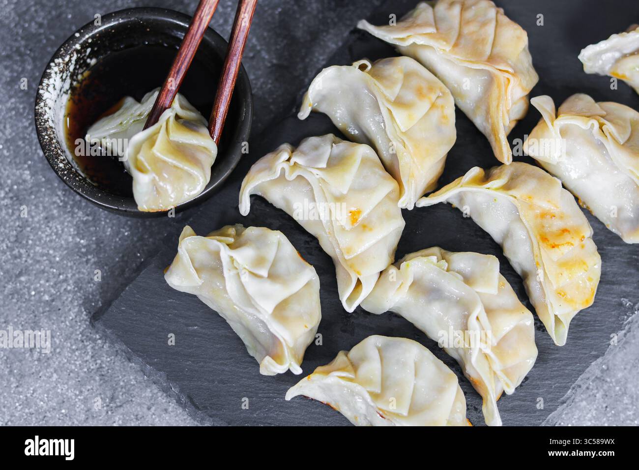 Ein Teller mit gekochten Gyoza-Knödeln, von denen einer mit Essstäbchen in eine kleine Schüssel Sojasauce getaucht wird, präsentiert auf einer dunklen strukturierten Oberfläche. Stockfoto