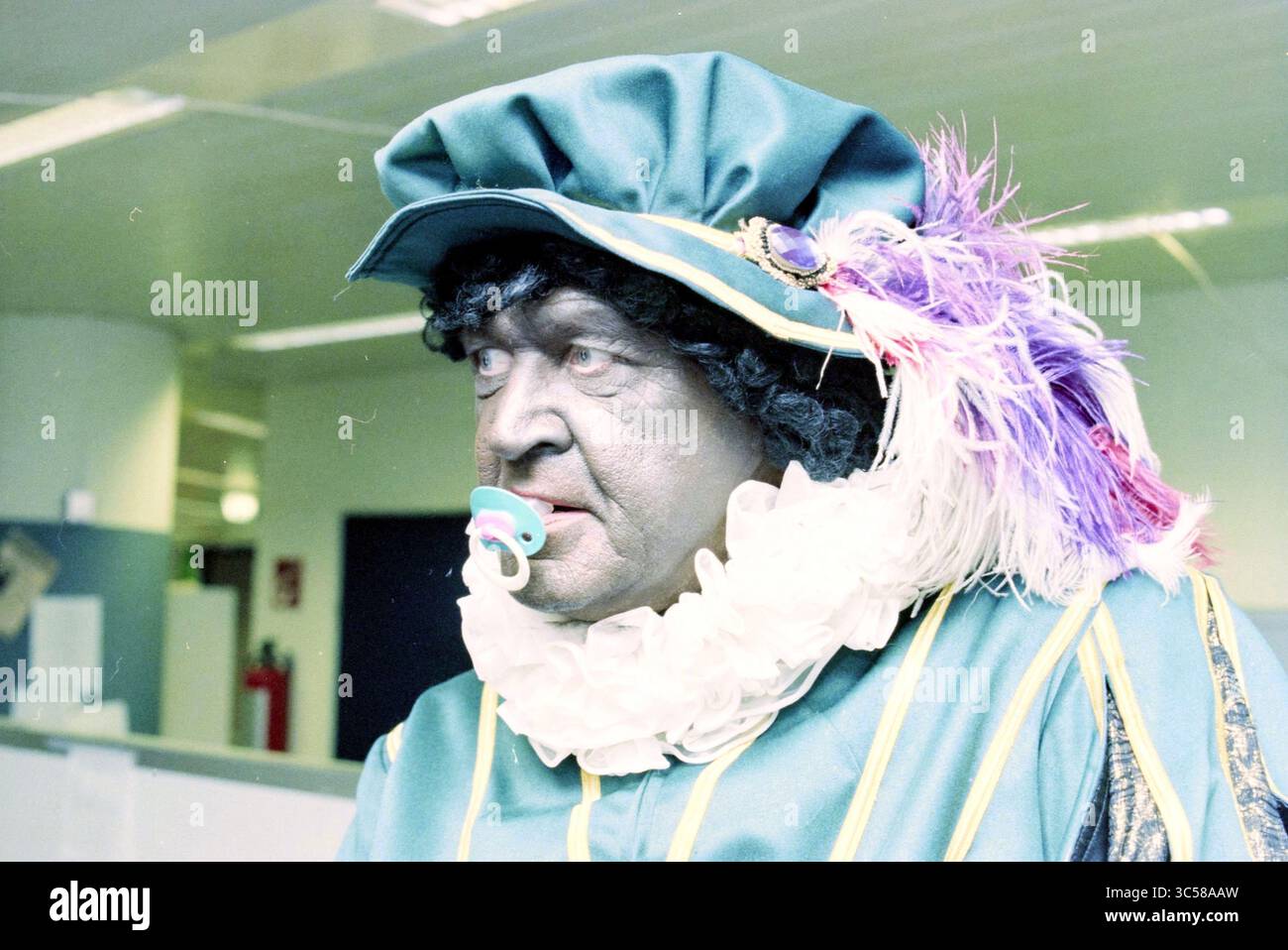 Black Pete, The Howler Whizgle News, Dutch Desk, Niederlande, 1950-2000 Eine Figur in einem bunten Kostüm, mit einem Rüschenkragen und einem dekorativen Hut mit Federn. Der Ausdruck ist ernst und kontrastiert mit der skurrilen Kleidung, während ein Schnuller eine unerwartete und humorvolle Note verleiht. Stockfoto