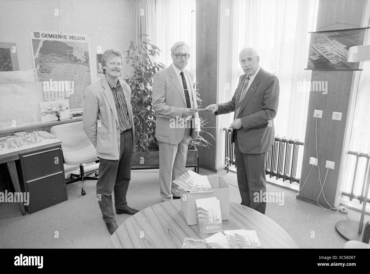 Präsentation des Mieterverbandes-Heftes. An Alderman Meyer, Books and Booksellers, Handing over and Transfers, 30-05-1989, Whizgle News, Dutch Desk, Niederlande, 1950-2000 drei Männer stehen in einem Büro, wobei einer den anderen ein Dokument vorlegt. Die Wände sind mit Plänen, Karten und Architekturmodellen verziert. Auf einem runden Tisch befindet sich eine Box mit Broschüren, die eine Diskussion oder Präsentation über ein Community-Projekt anzeigt. Stockfoto